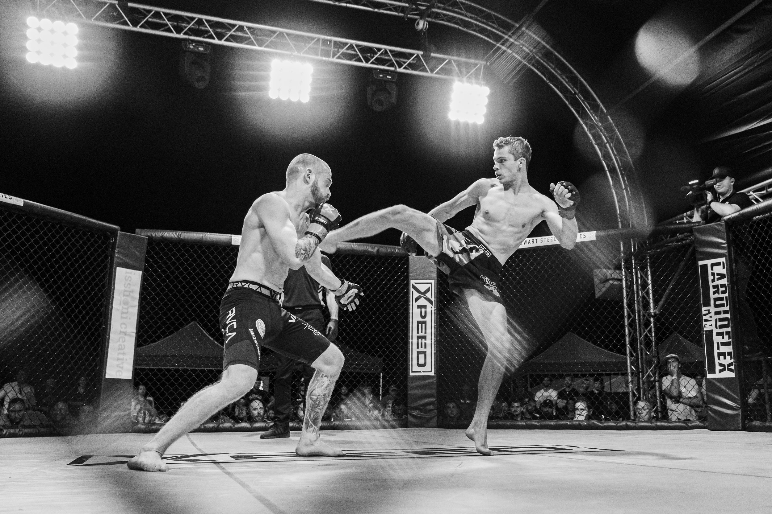 Two male mixed martial artists fighting inside an octagon cage, one delivering a high kick to the other's head, with a referee nearby, under bright overhead lights, in an arena with spectators.