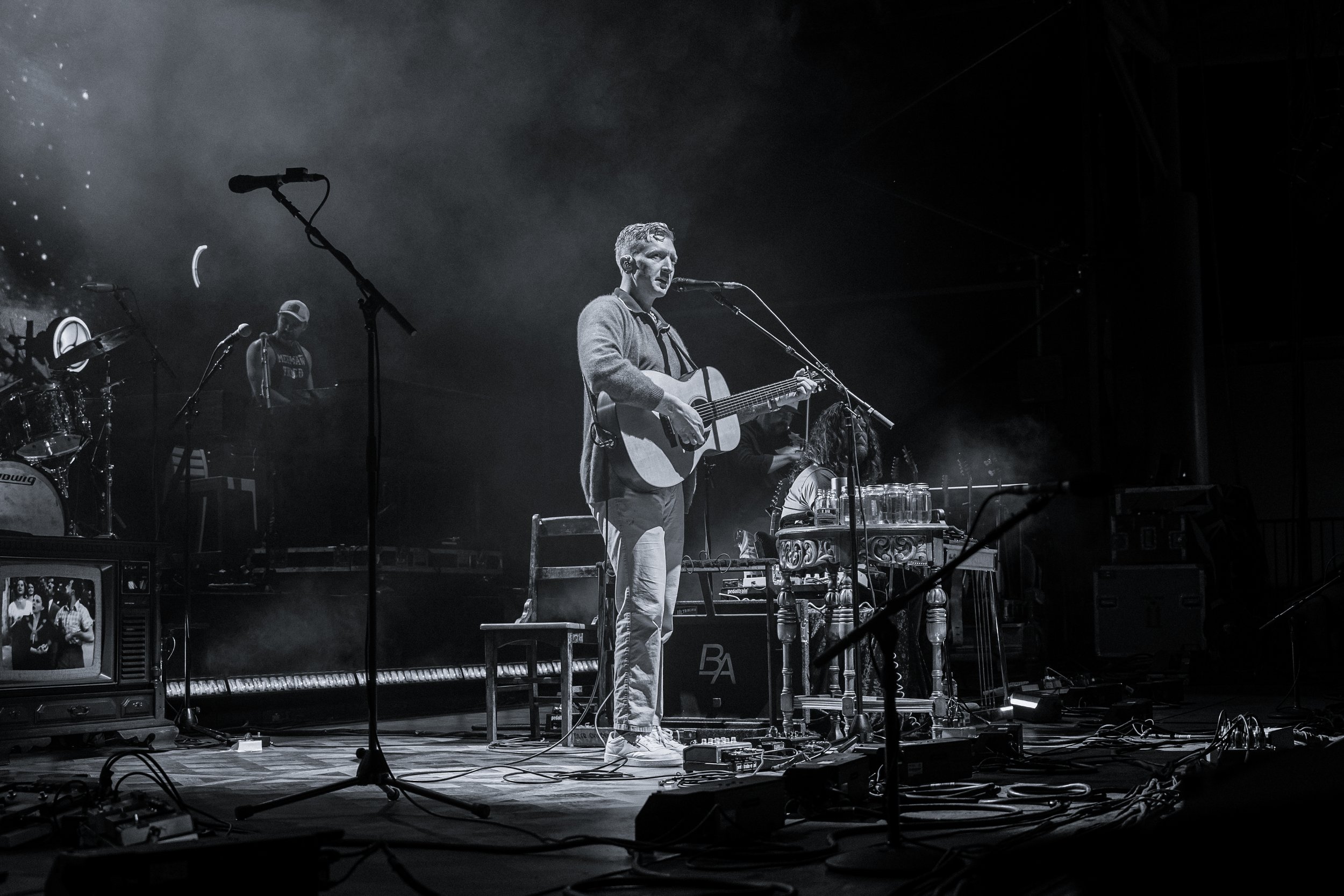 A black and white photo of a man playing an acoustic guitar and singing into a microphone on a stage. There are other band members and musical equipment visible in the background.