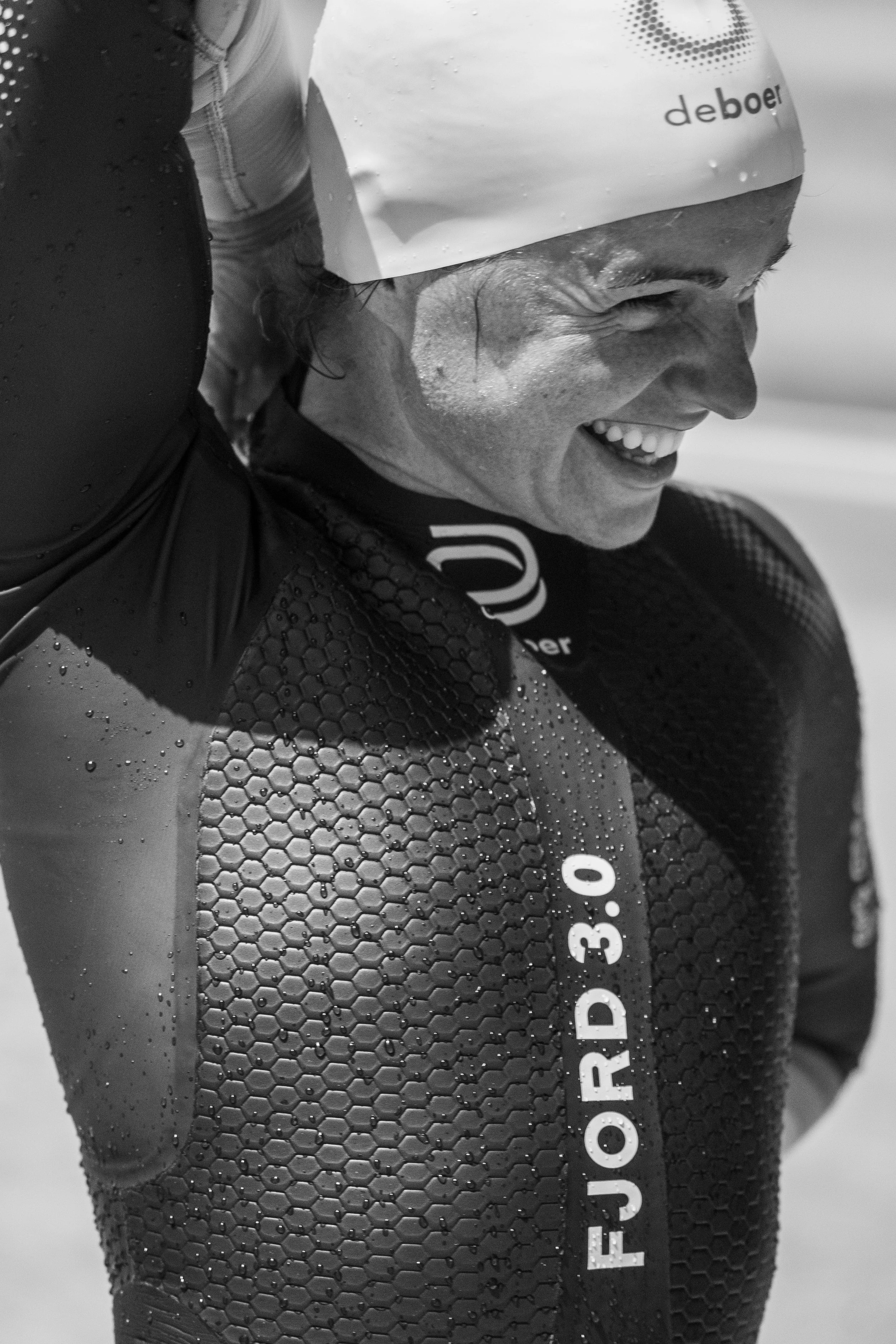 Close-up of a smiling athlete wearing a swim cap with water droplets, in a wetsuit with a honeycomb pattern, and a black and white background.