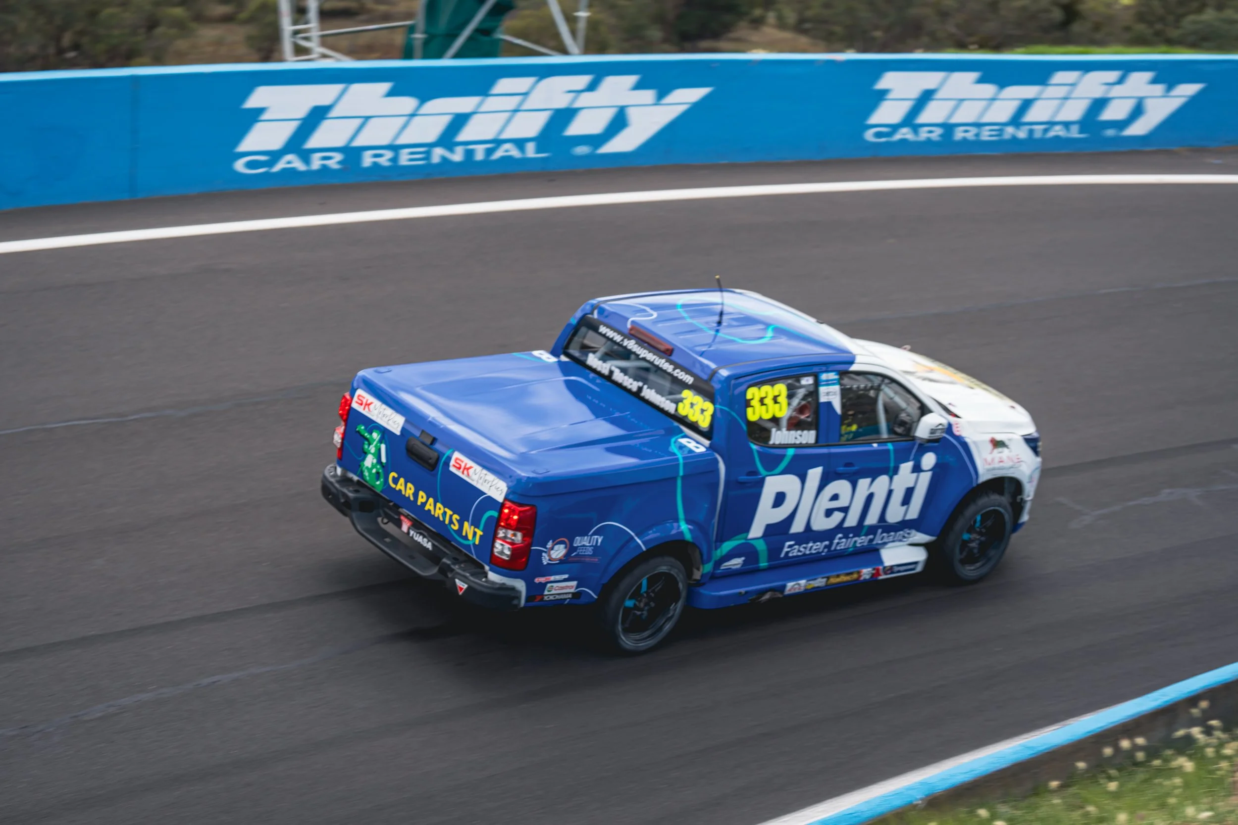A blue race truck with the number 333 on the side, branded with Plenti and various sponsor logos, driving on a racing track with a blue barrier advertising Thrifty Car Rental.
