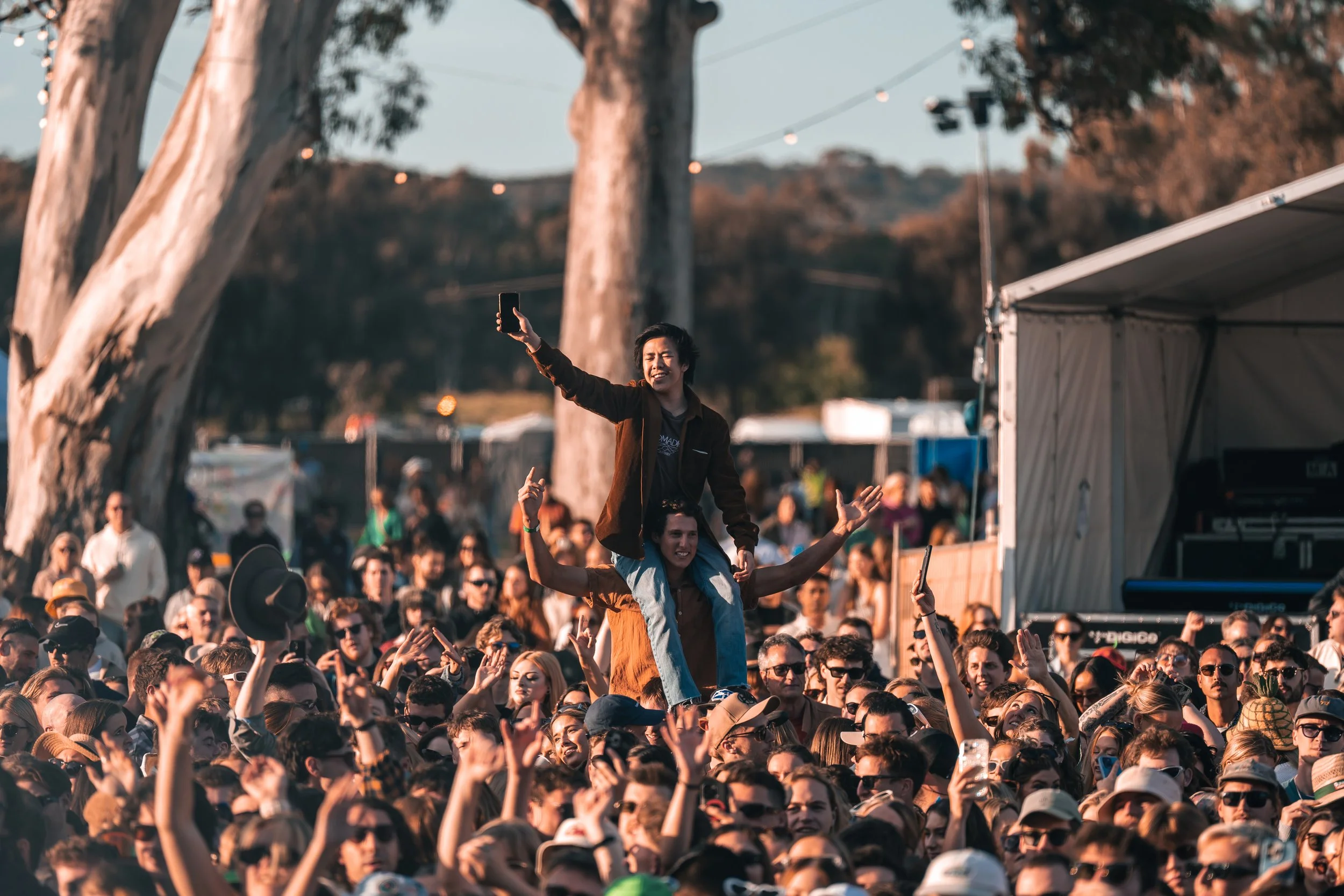 A large crowd at an outdoor event with two women in the center, one sitting on the other's shoulders, taking a selfie. The crowd is engaged, and there are trees and string lights overhead.