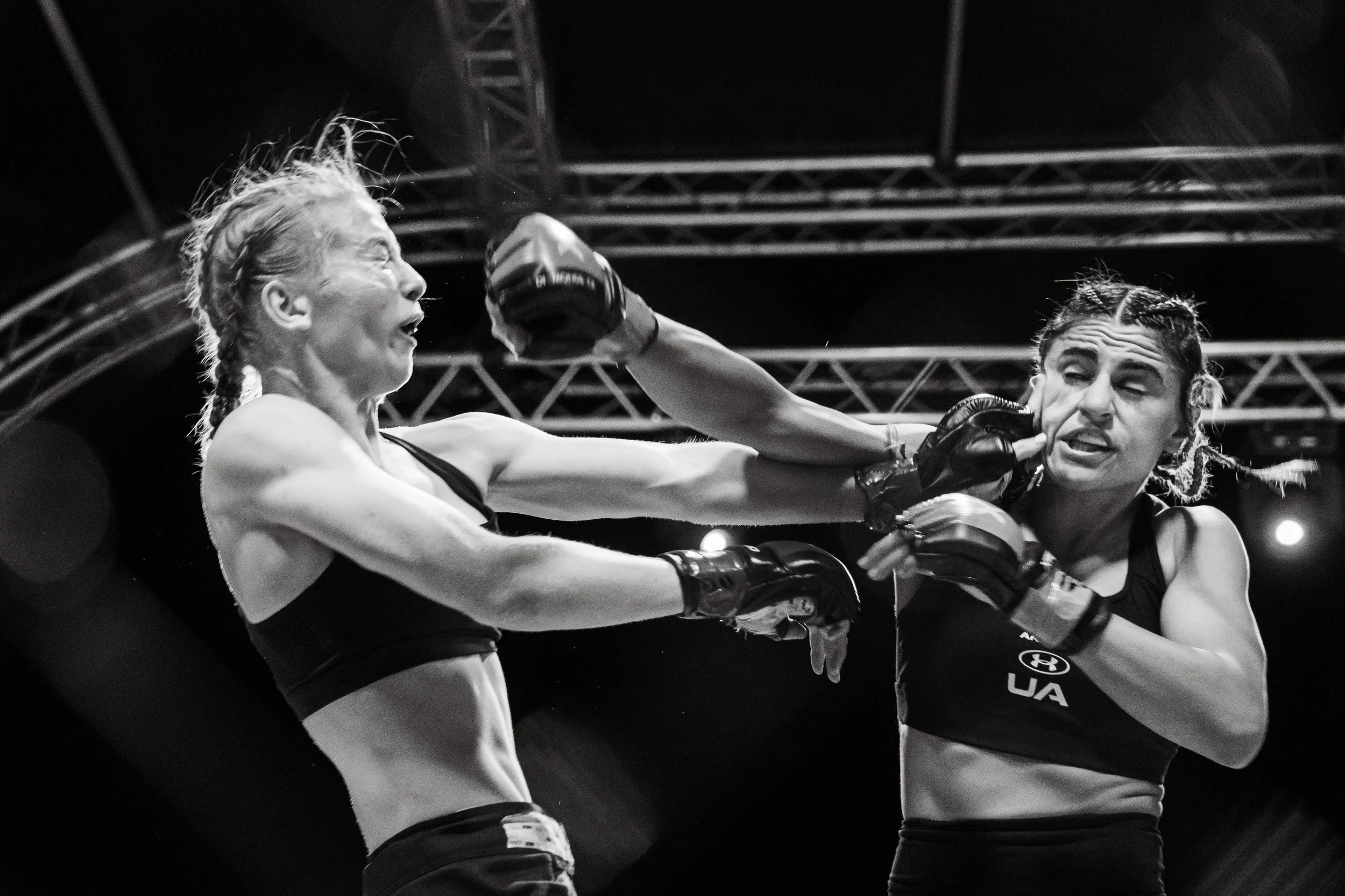 Two female mixed martial artists are fighting in a ring, with one delivering a punch to the other. The scene is intense, set under bright lights with metal structures visible in the background.