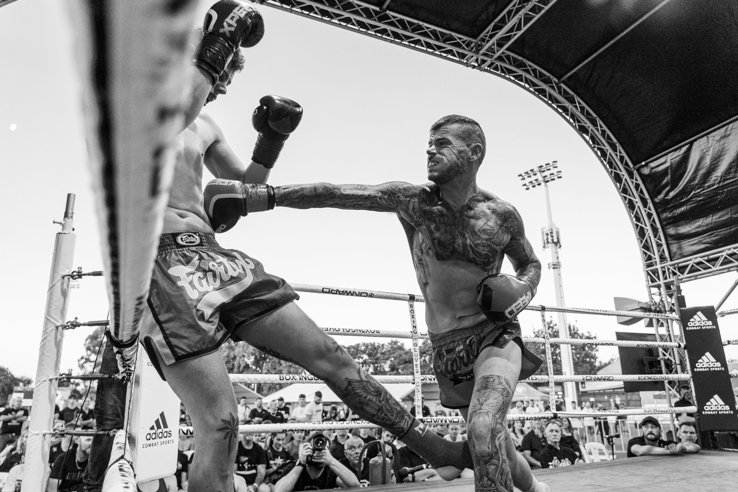 A black and white photo of two male fighters in a boxing ring during a match. One fighter is punchiing the other, who is on the ropes. Spectators and officials are visible outside the ring, and there is a large arched canopy overhead.