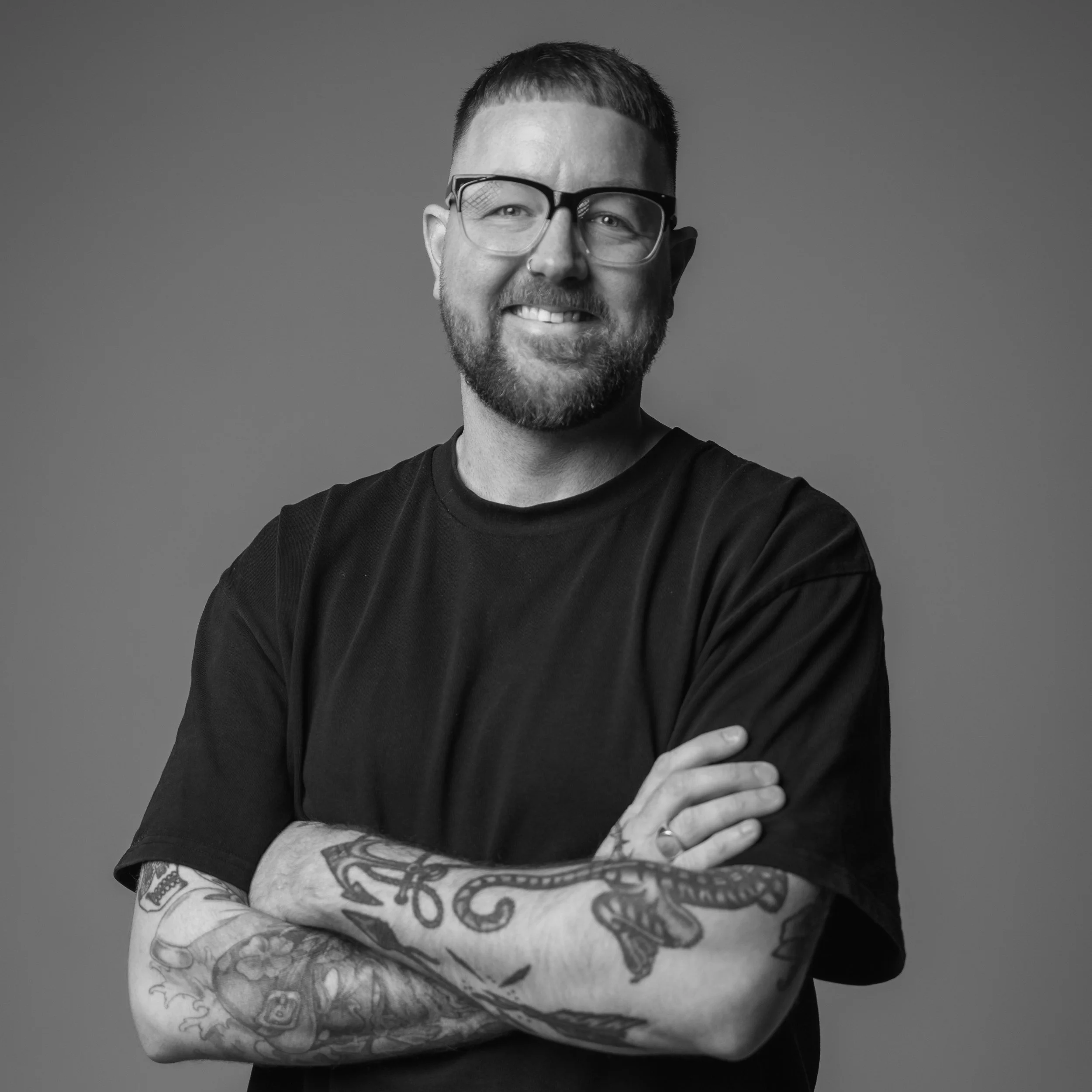 Black-and-white photo of a man smiling, wearing glasses, with tattoos on his left arm, crossed arms, and short hair.