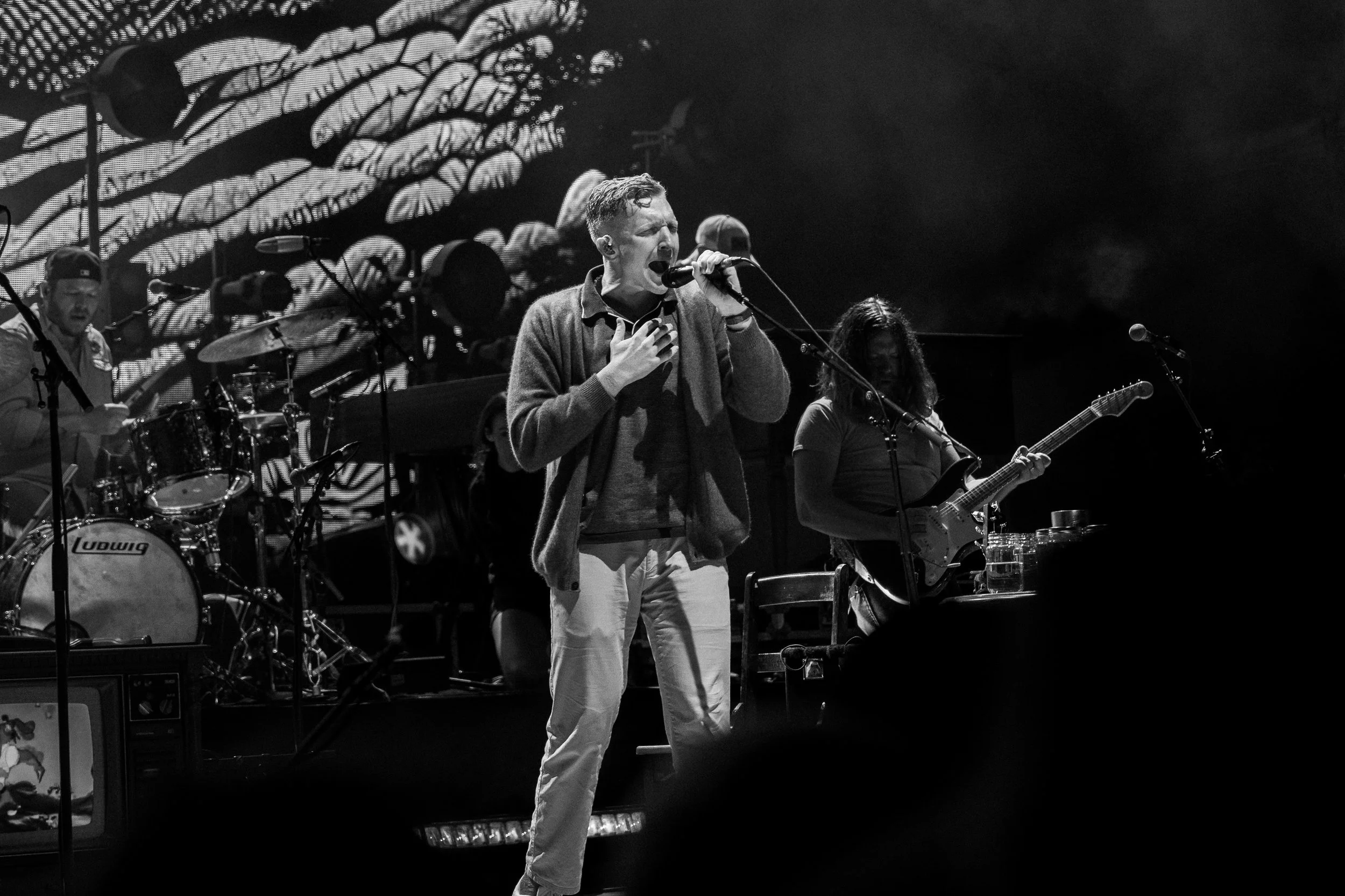A live musical performance featuring a male singer with short hair and a sweater, singing passionately into a microphone, accompanied by a guitarist and a drummer, on a stage with lighting and visual effects in the background.