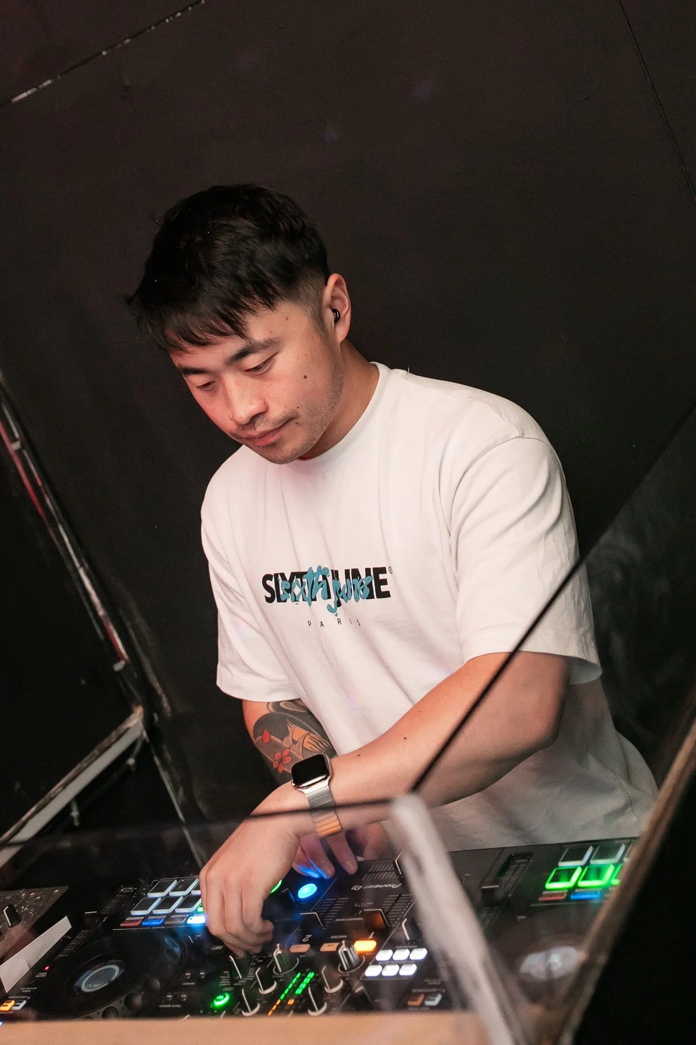 A DJ with dark hair, wearing a white T-shirt with text, is mixing music on a DJ controller with colorful lights. The background is dark.