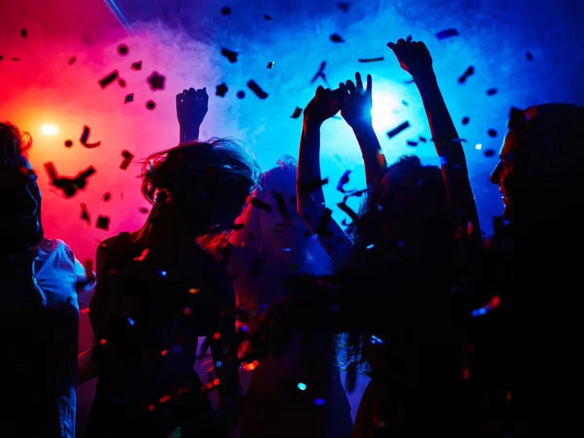 People dancing and celebrating with confetti in a dark room with colorful lights.