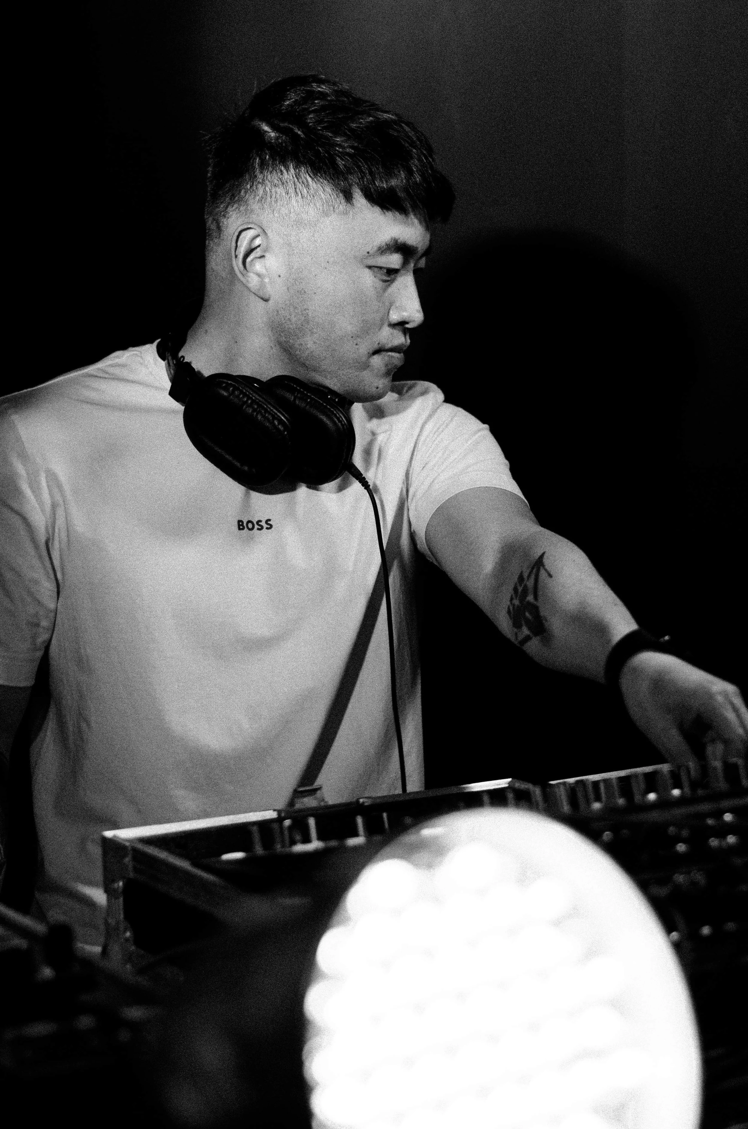 A black and white photo of a young man with short dark hair and a fade haircut, wearing headphones around his neck and a white t-shirt with the word "BOSS" on it. He appears to be DJing or working with audio equipment, extending his arm towards a mixer or turntable.