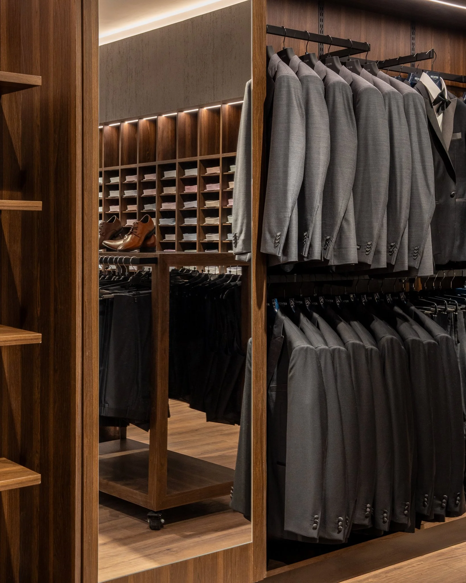 Interior of a clothing store featuring gray suits hanging on racks and brown leather shoes displayed on a shelf, with wood and dark finishes.