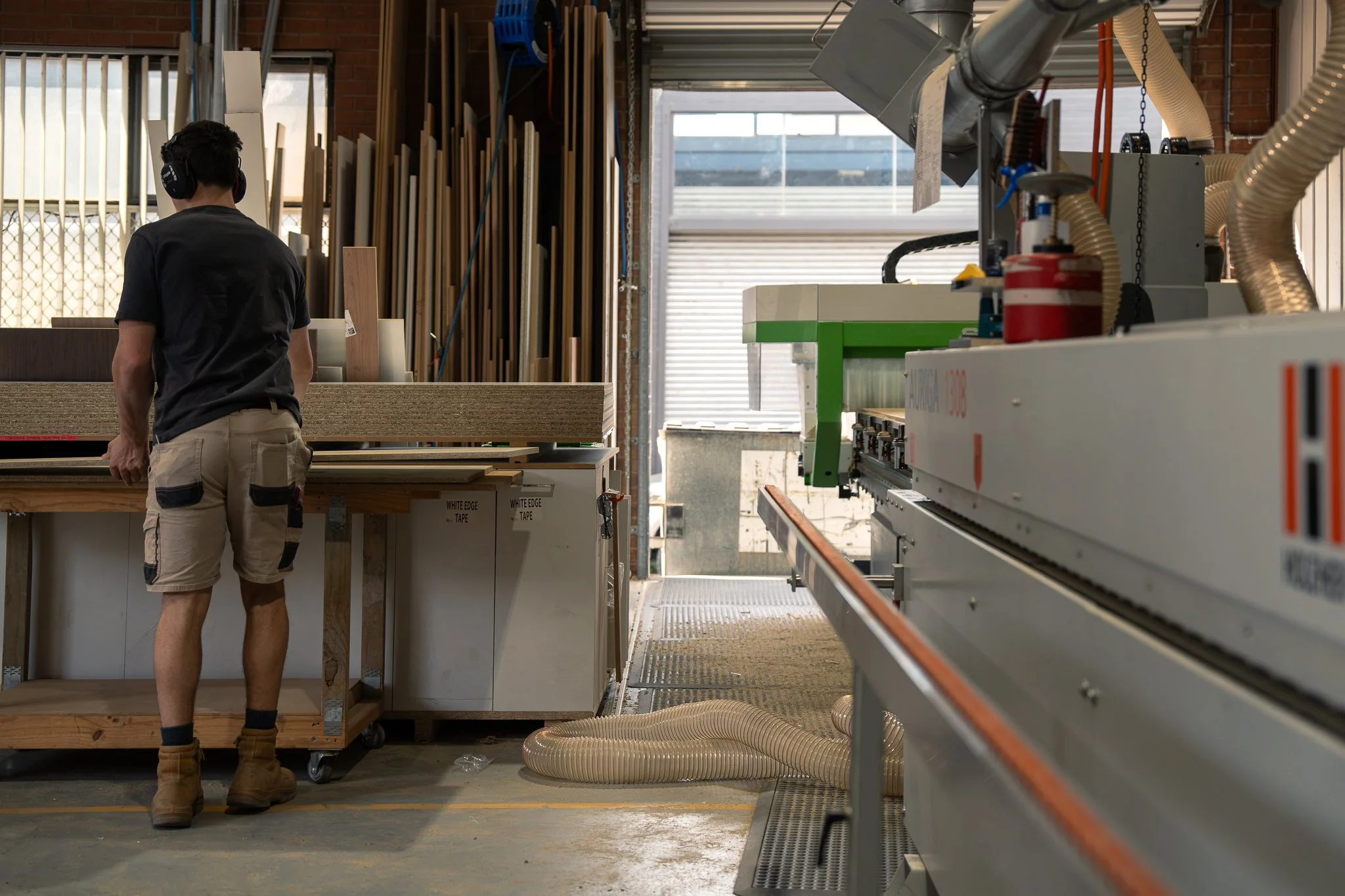 A worker in a woodworking shop wearing ear protection, standing at a workbench with wood panels, surrounded by woodworking machinery and materials.
