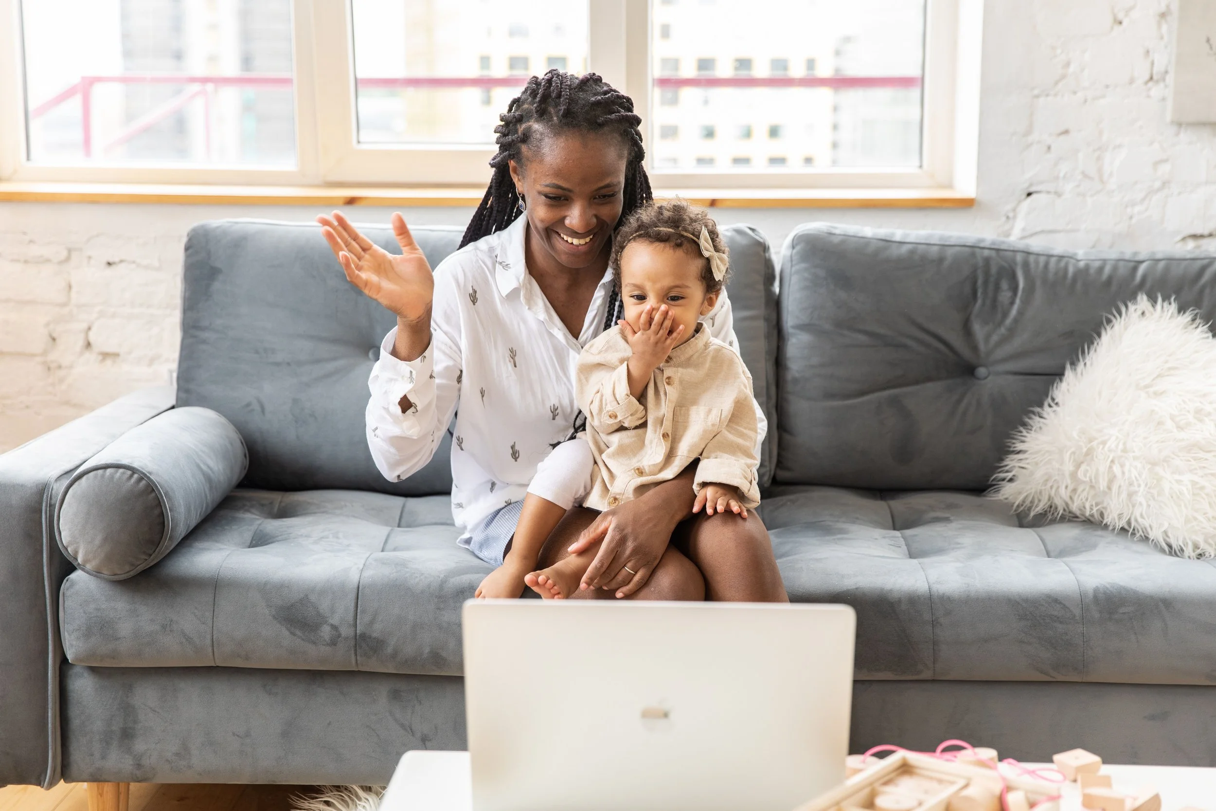 A woman and a young girl sitting on a gray couch in a bright room, watching something on a laptop, with their hands raised and smiling.