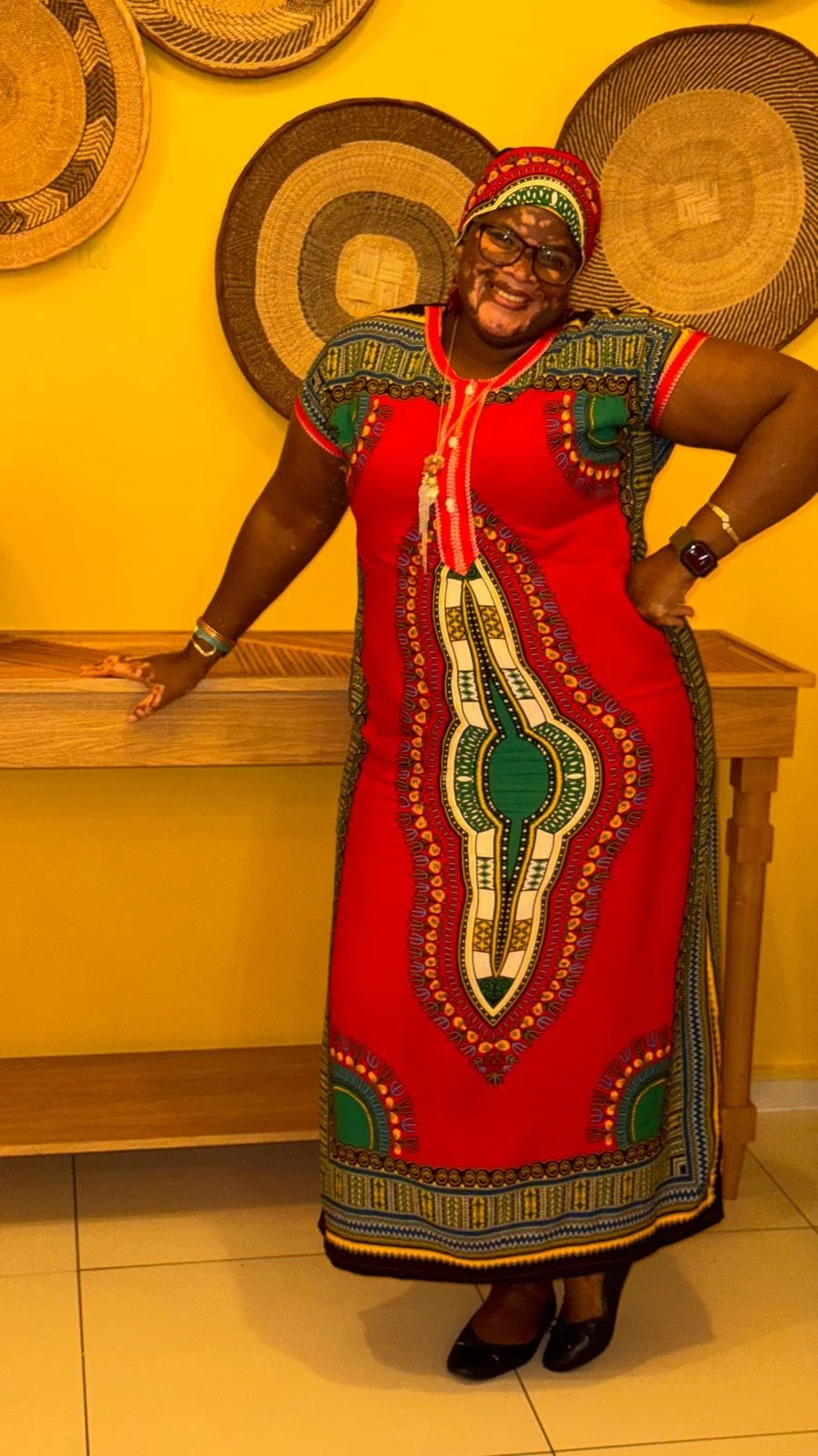 A person wearing traditional African attire, including a red dress with intricate green, yellow, and white patterns, standing in front of a yellow wall with woven baskets.
