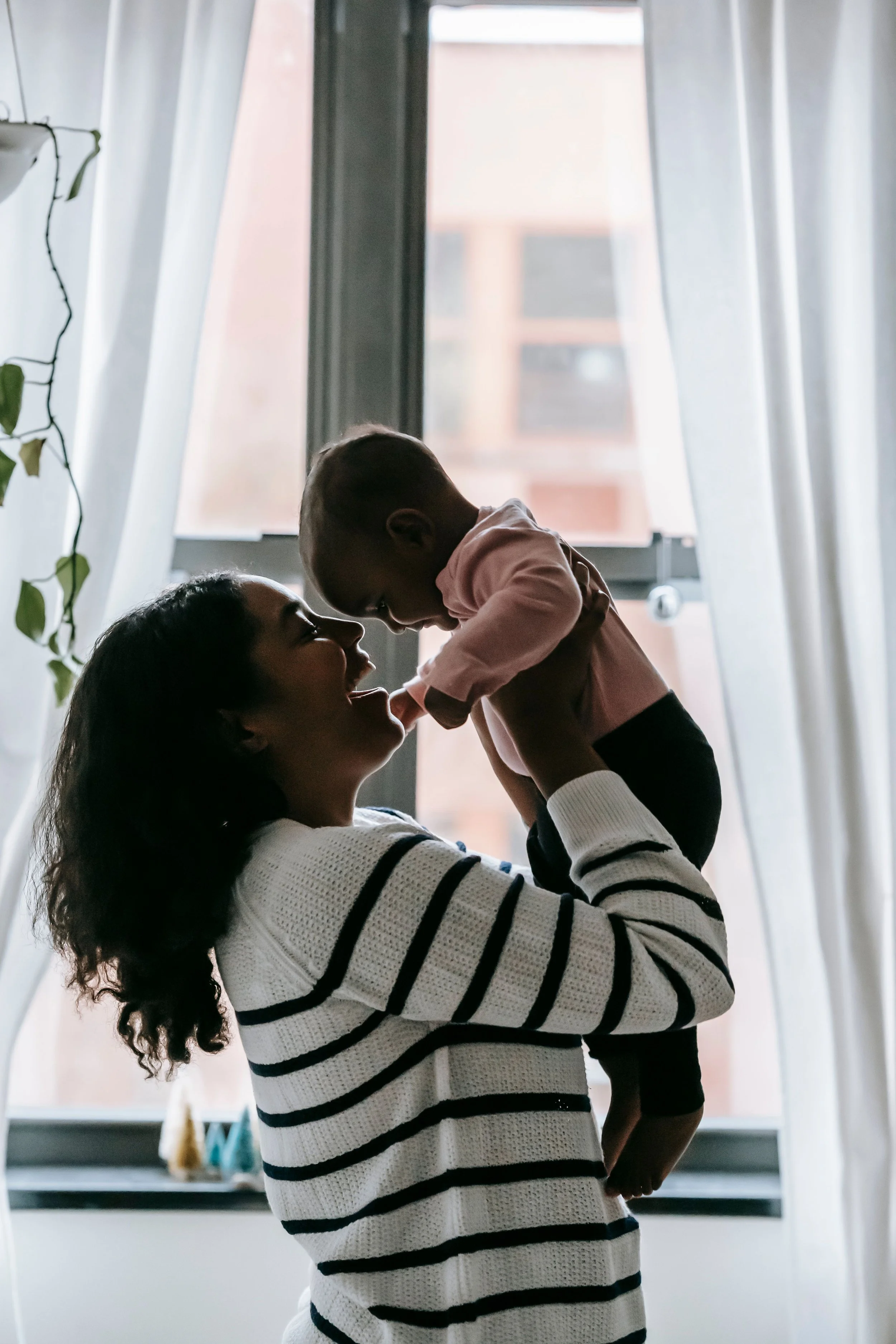 A woman holding a young boy up in front of a window with sun-drenched curtains. They are smiling and facing each other, sharing a joyful moment indoors.