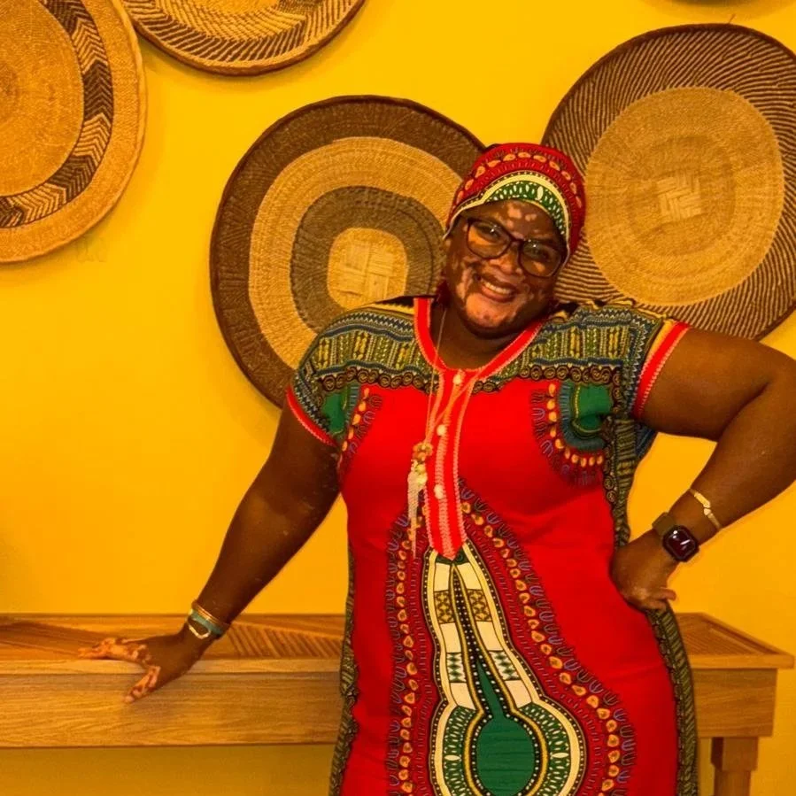 A smiling woman wearing glasses and a colorful African-style dress, standing in front of a yellow wall with woven baskets hanging on it.