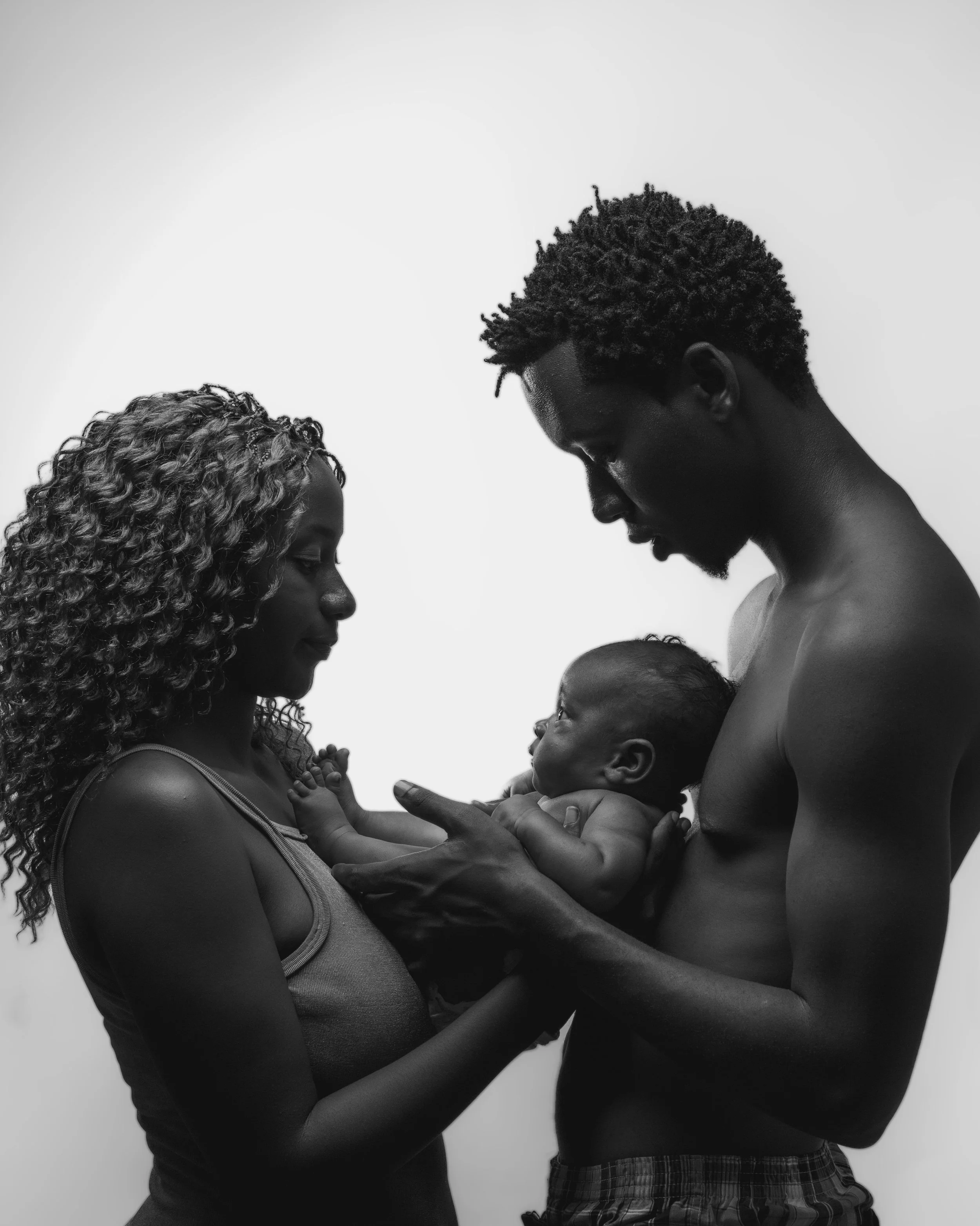 A black and white photo of a man and woman looking at each other, holding a baby between them, with the woman and man both touching the baby's hands.