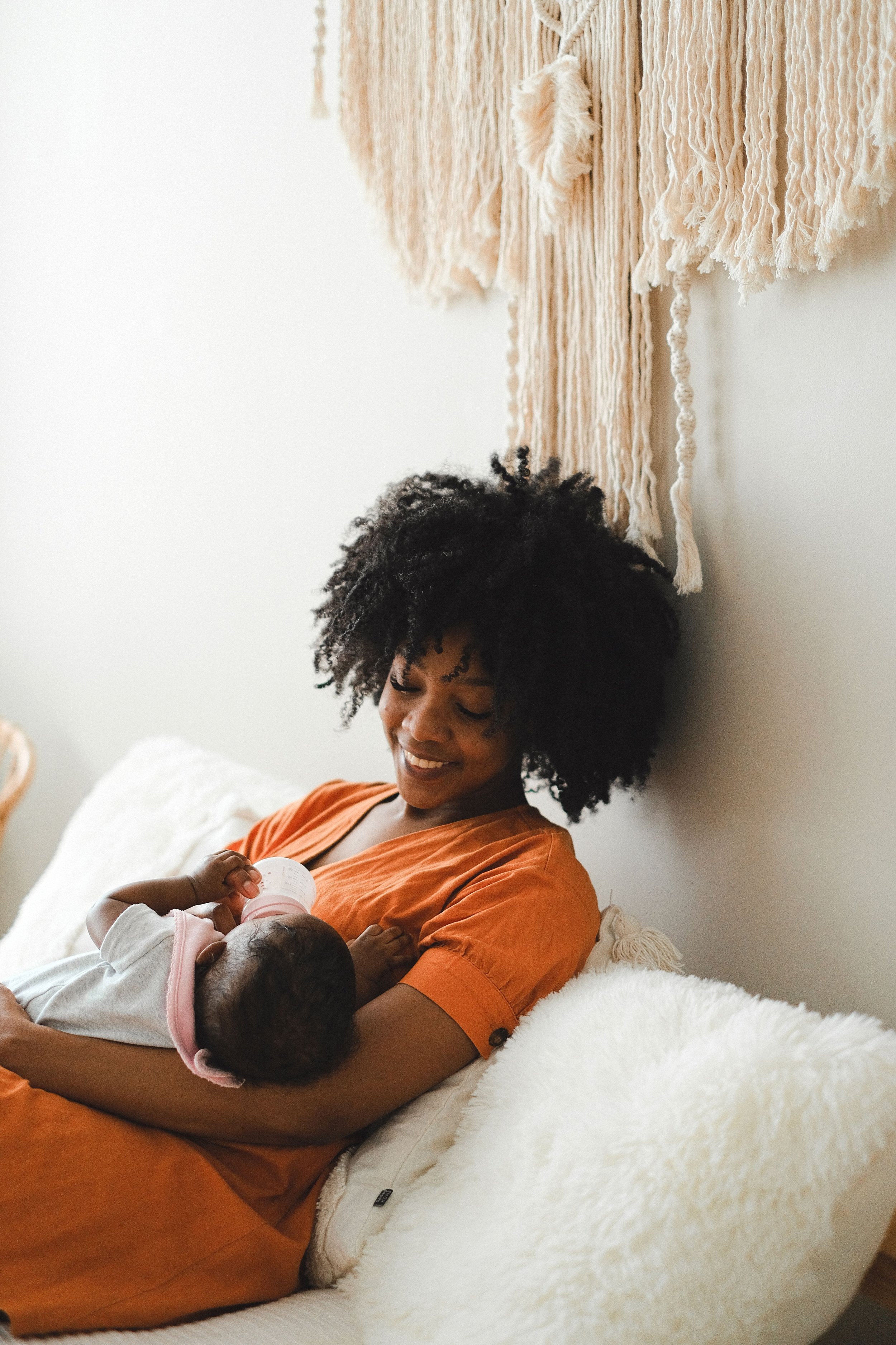 A woman with curly black hair smiling while holding two babies, one drinking from a bottle, on a white plush couch in a room with neutral decor and a beige wall hanging.