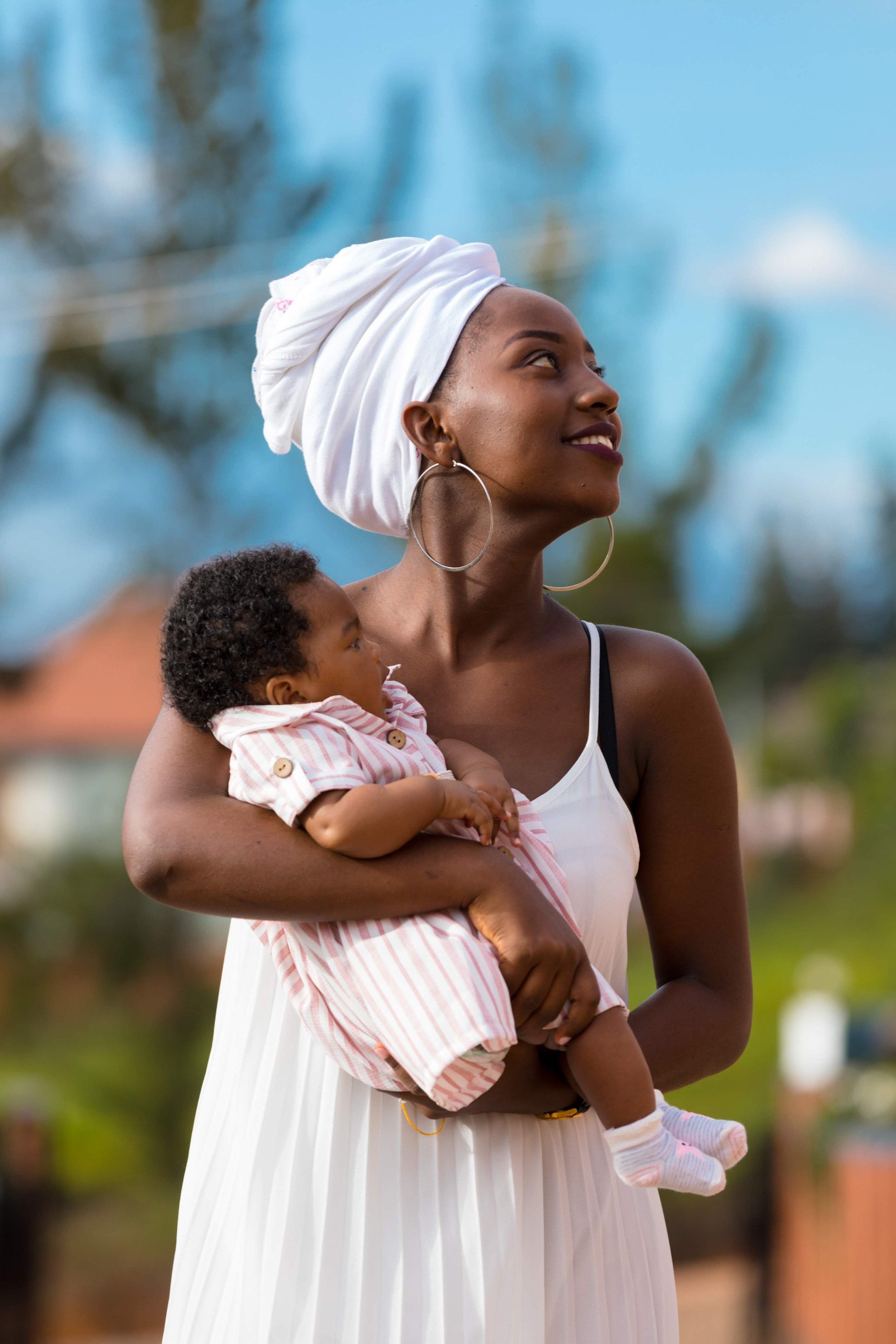 A woman with dark skin and earrings holds a small child in her arms outdoors, both smiling with trees and blue sky in the background.