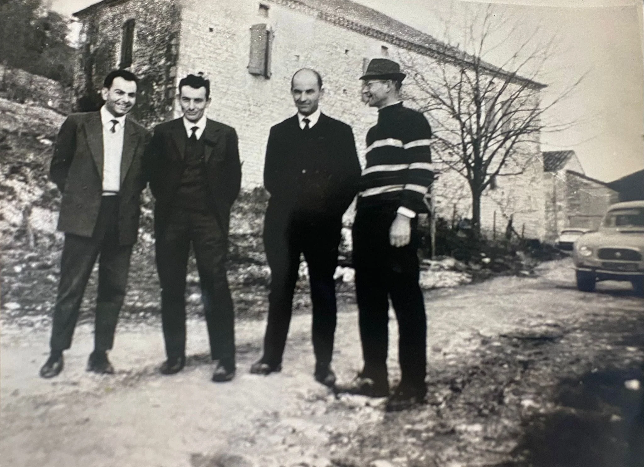 Four men standing outdoors on a snowy day, with a large tree and buildings in the background. Three are dressed in suits, one in a striped sweater and hat.