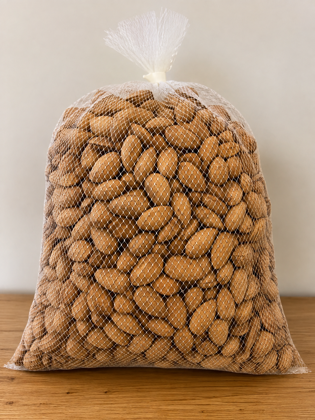 A large, clear plastic net bag filled with unshelled peanuts placed on a wooden surface against a plain beige wall.