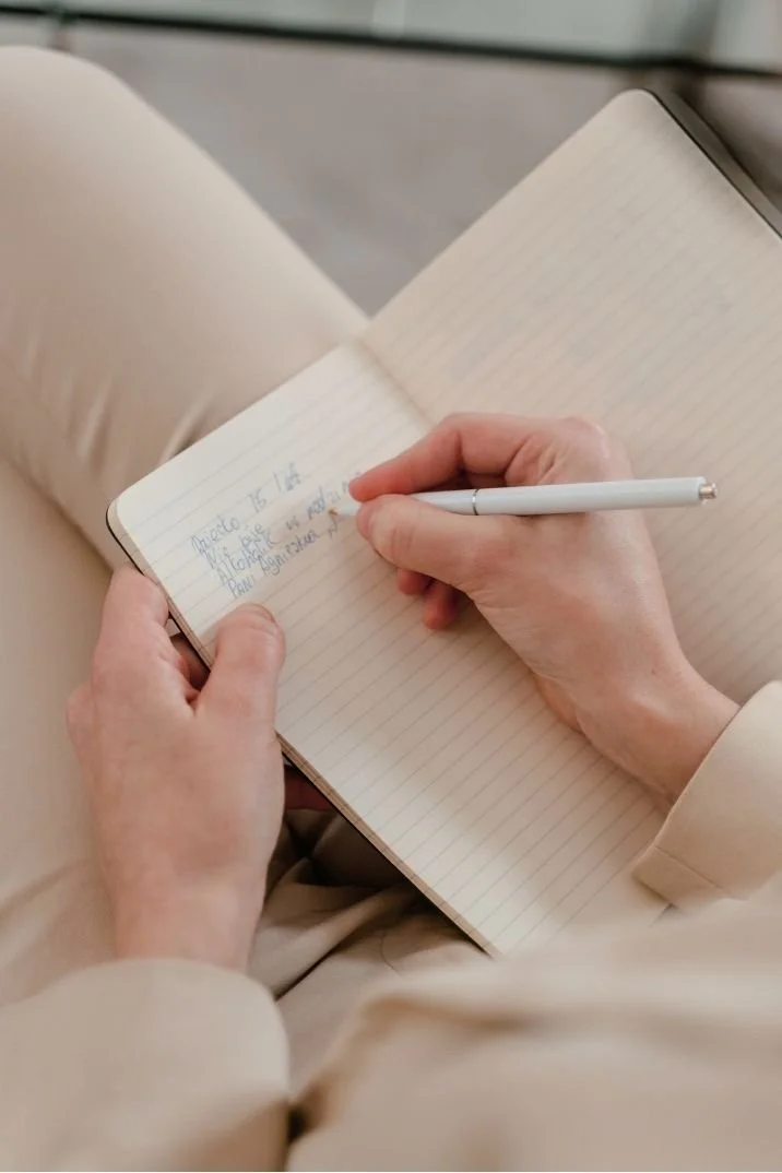 A person writing in a notebook with a white pen while sitting with legs crossed, wearing light-colored clothing.