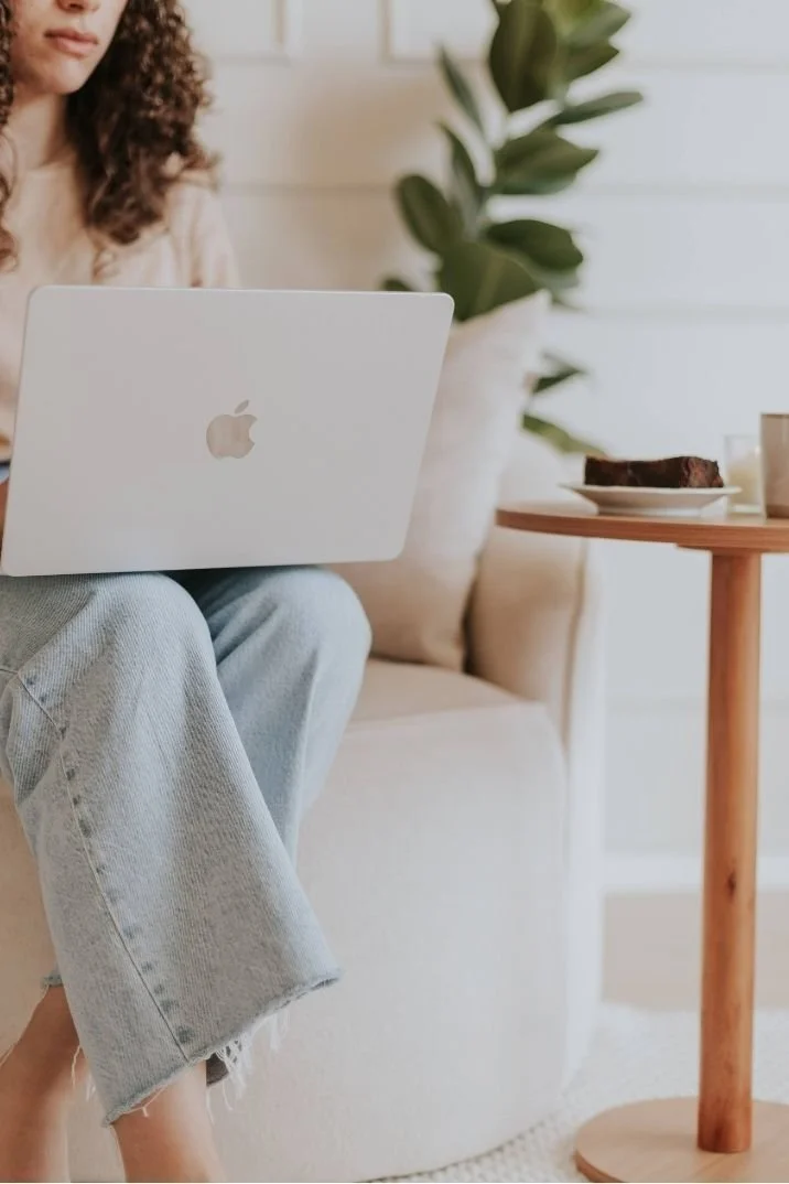 A person with curly hair working on a white MacBook laptop in a cozy, well-lit living room with a large green plant in the background. They are sitting on a beige sofa with a small wooden table next to them, which has a plate with a piece of cake and some cups.