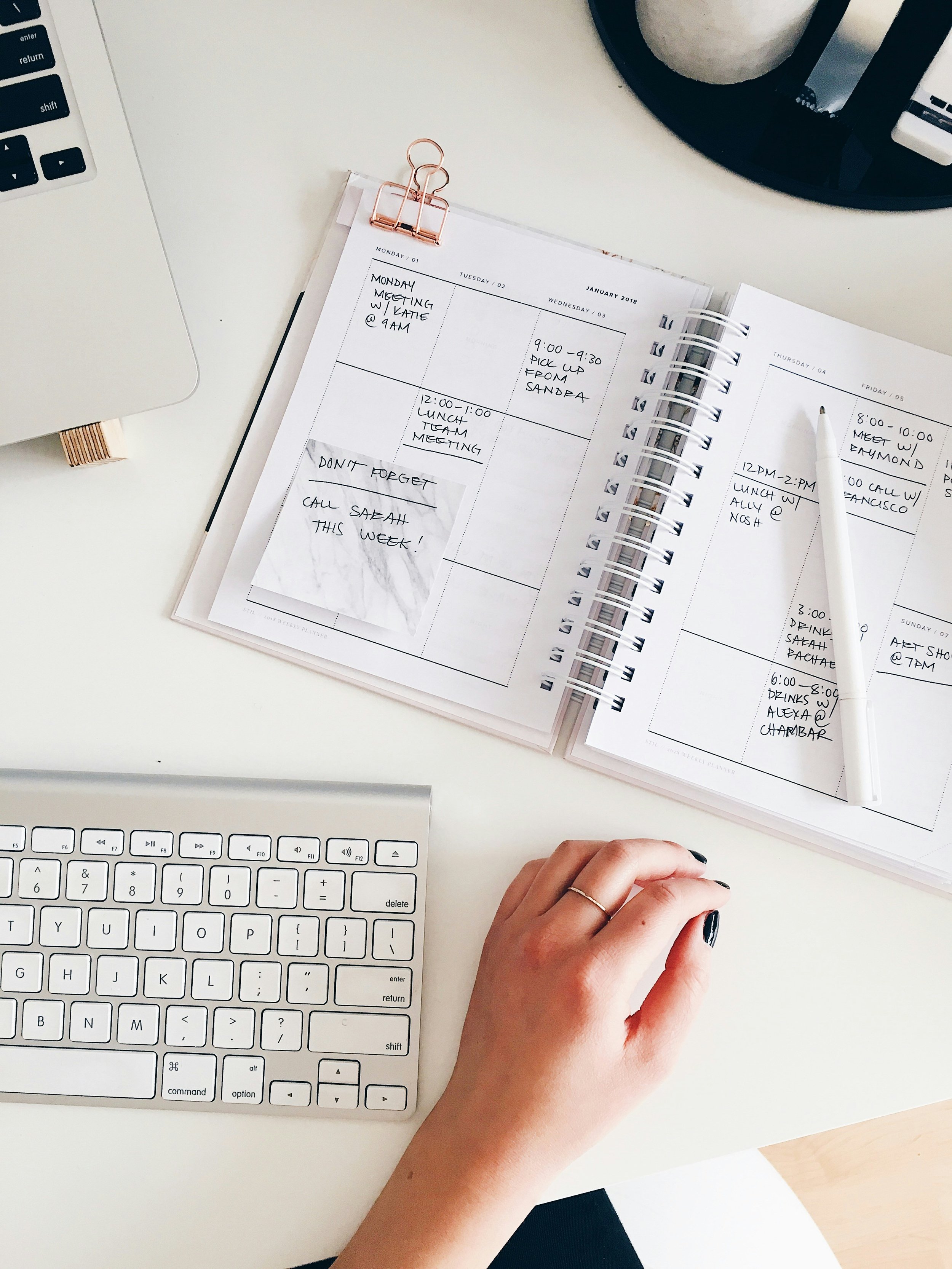 A workspace with a white computer keyboard, a computer mouse, a planner open with handwritten notes and appointments, a white pen, and a person's hand with black painted nails resting on the desk.