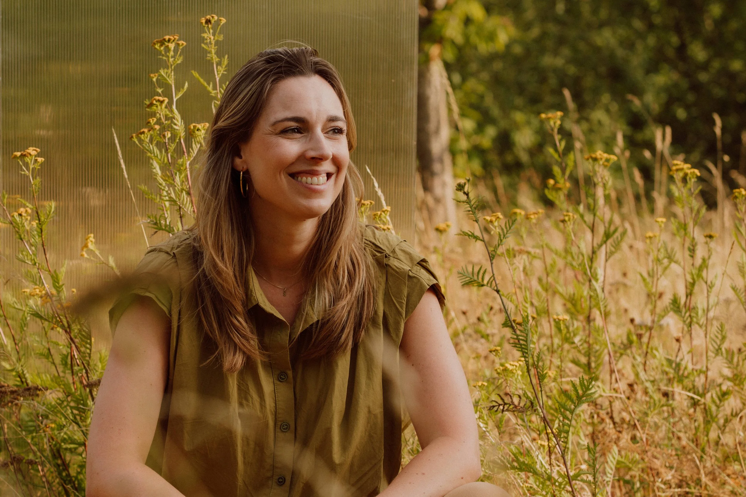 Vrouw met lang, bruin haar die in een veld met gele bloemen zit, glimlacht en kijkt naar rechts.