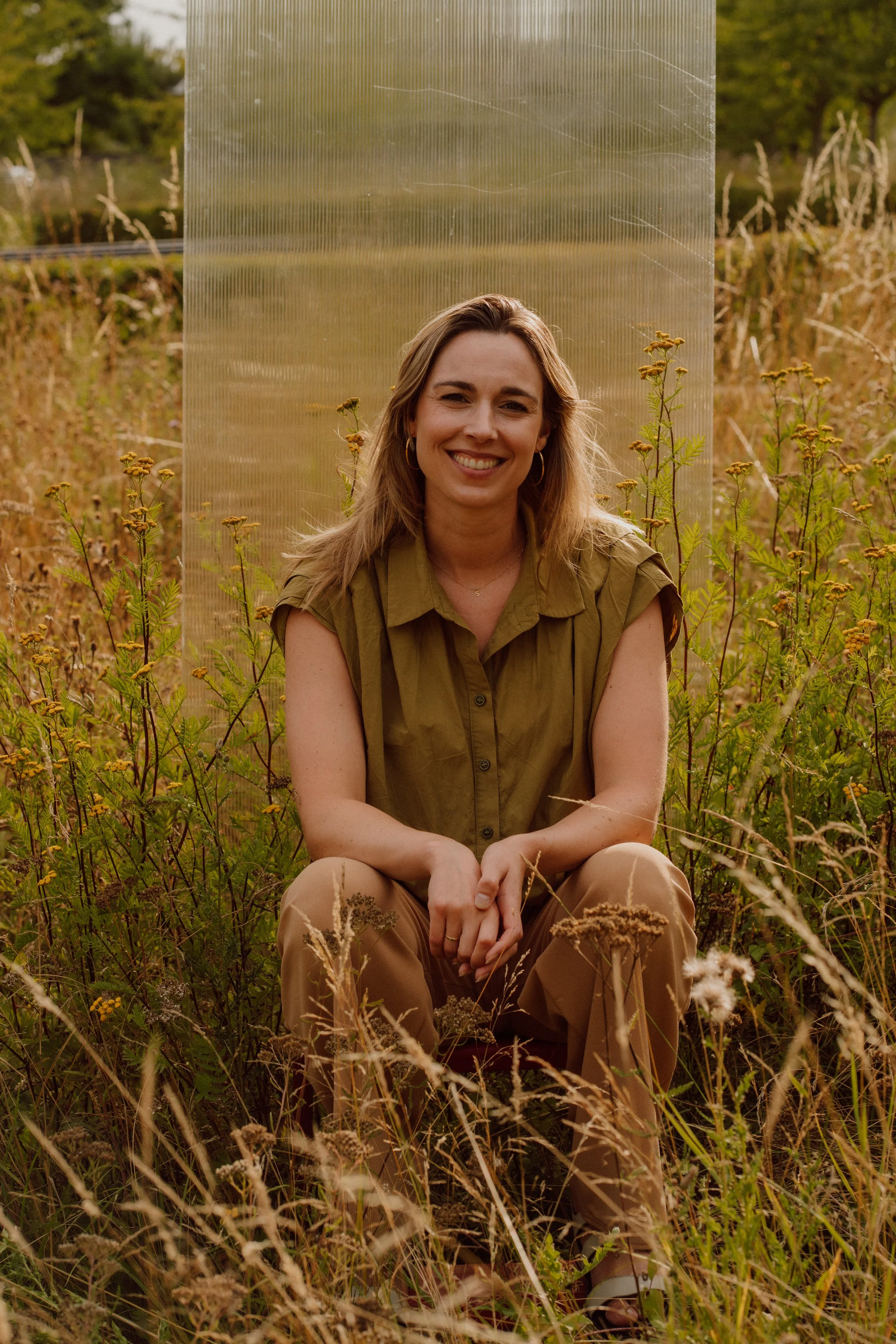 Vrouw zittend in een veld met gele bloemen, lachend naar de camera, tijdens zonsondergang of schemering.