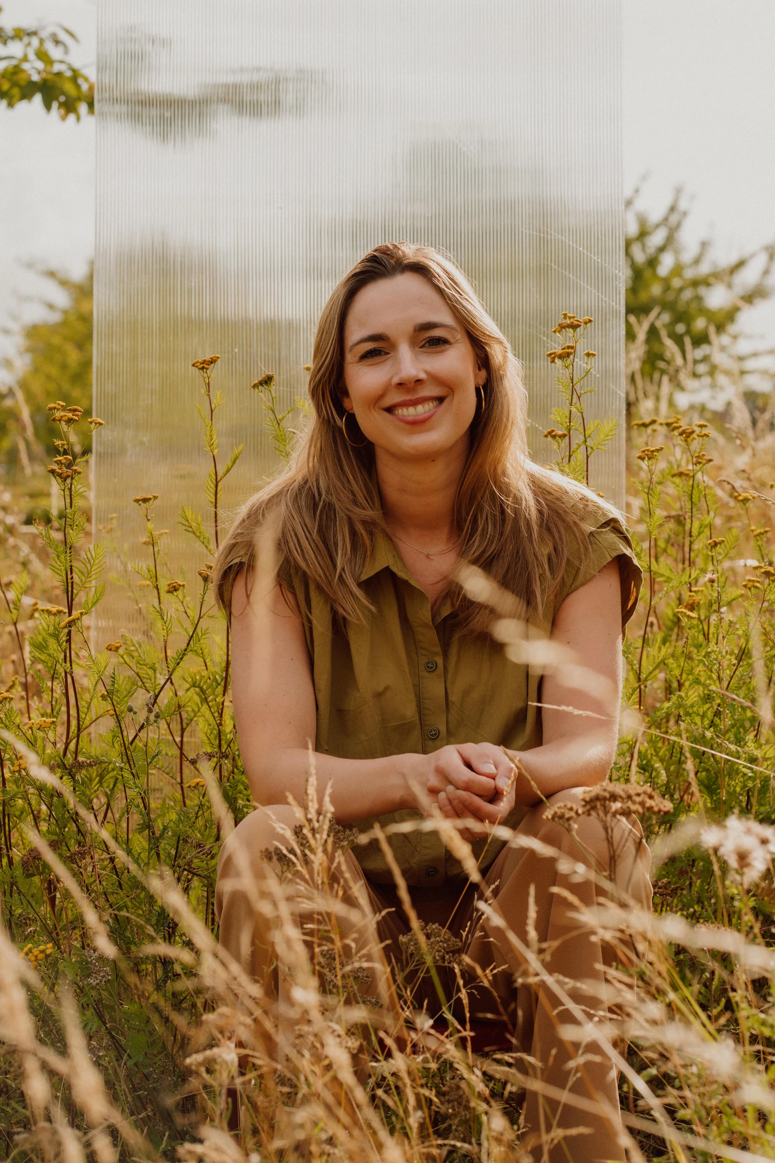 Vrouw in een veld met grassen en bloemen, zittend voor een plastic scherm, glimlachend.