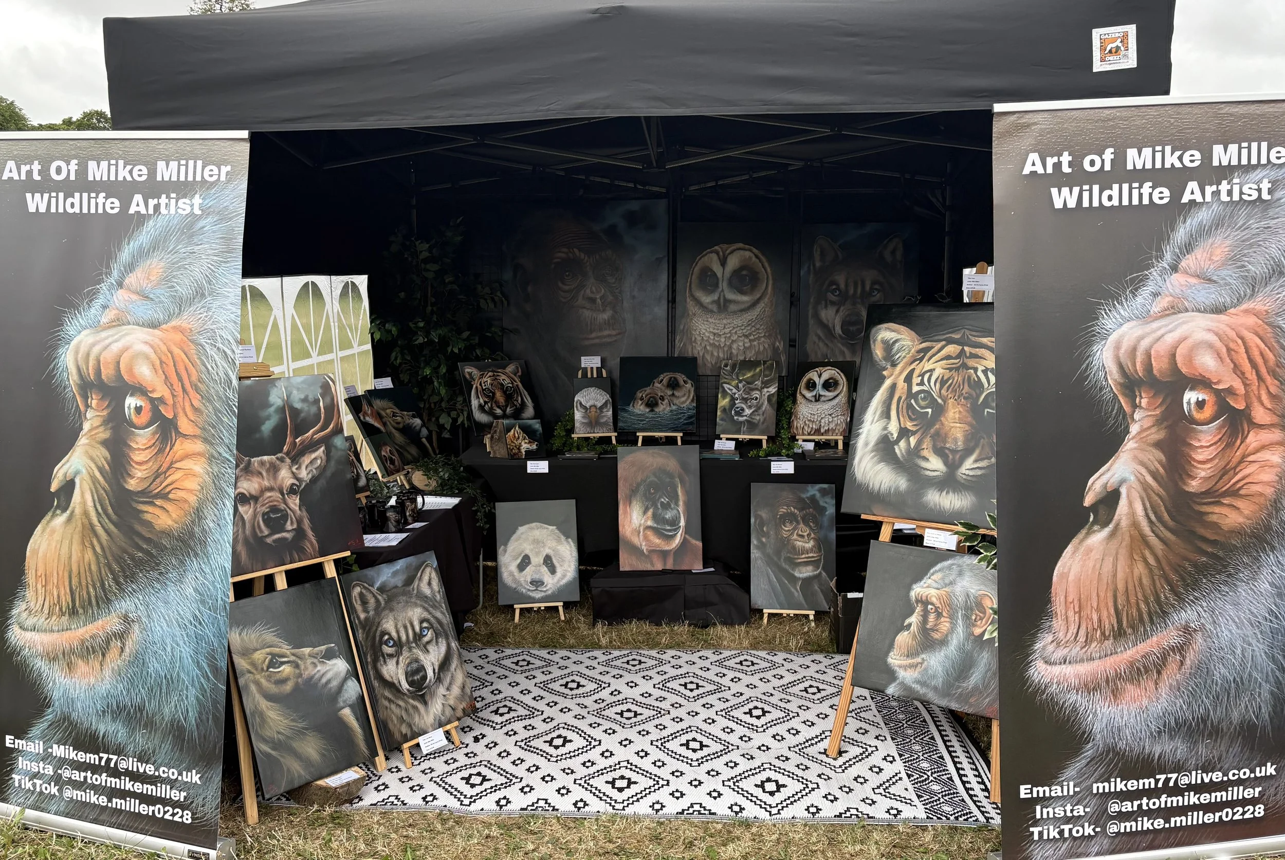 Art display booth showcasing wildlife paintings by Mike Miller, including portraits of a lion, wolf, owl, bear, jaguar, and other animals, with contact information on banners.