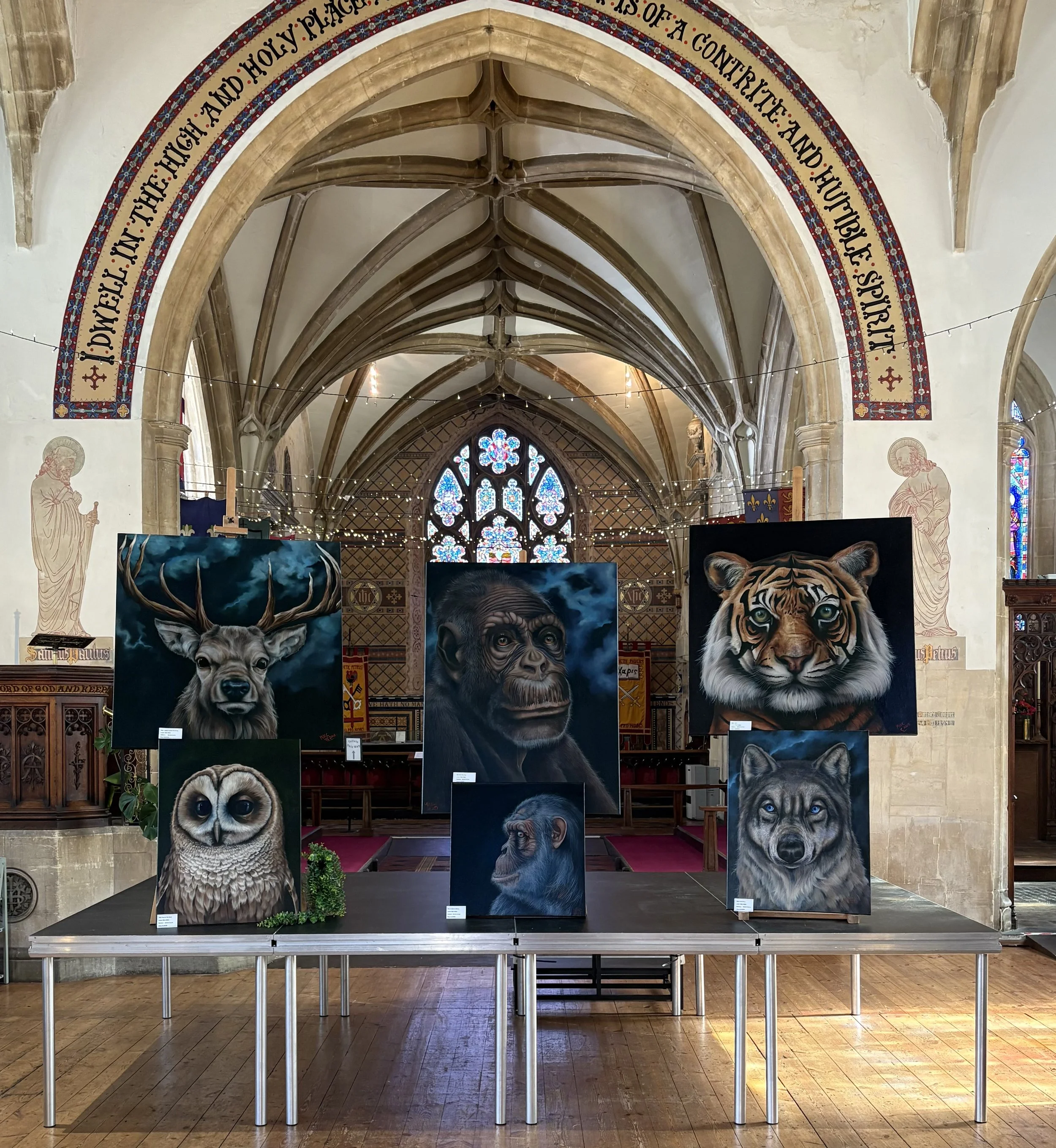 Art display featuring realistic paintings of various animals including a deer, a gorilla, a tiger, an owl, a wolf, and a dog, set inside a church with stained glass windows and ornate architectural details.