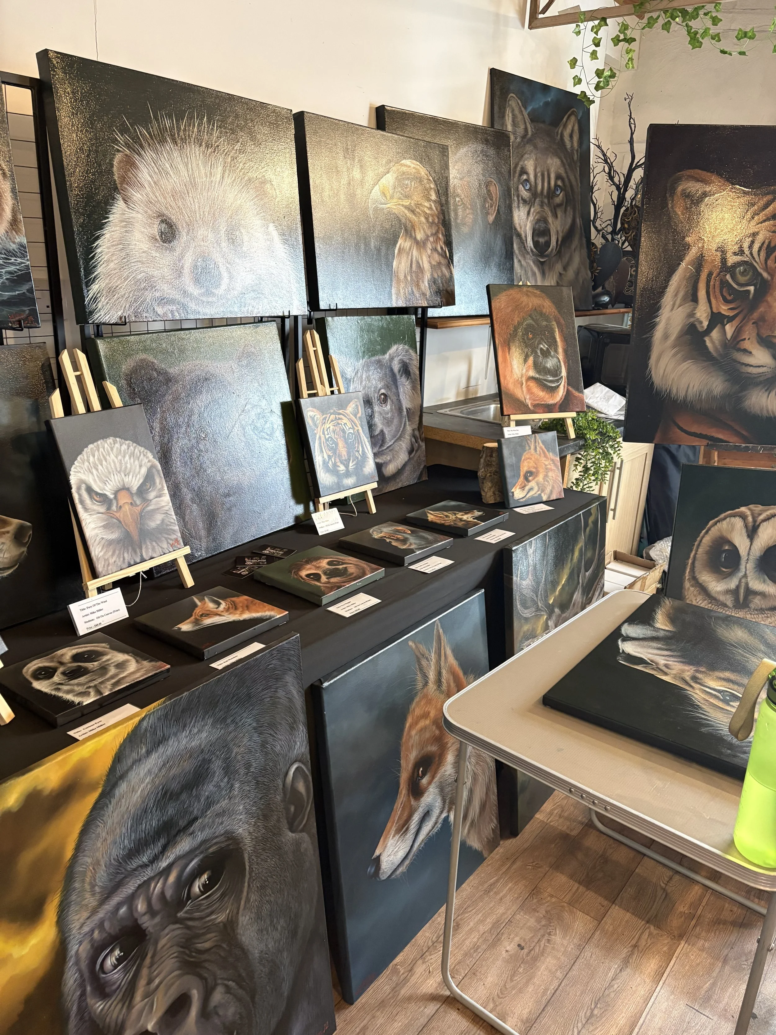 Wall display of animal paintings, including a monkey, eagle, wolf, tiger, and bear, with various paintings on tables and easels in a gallery setting.