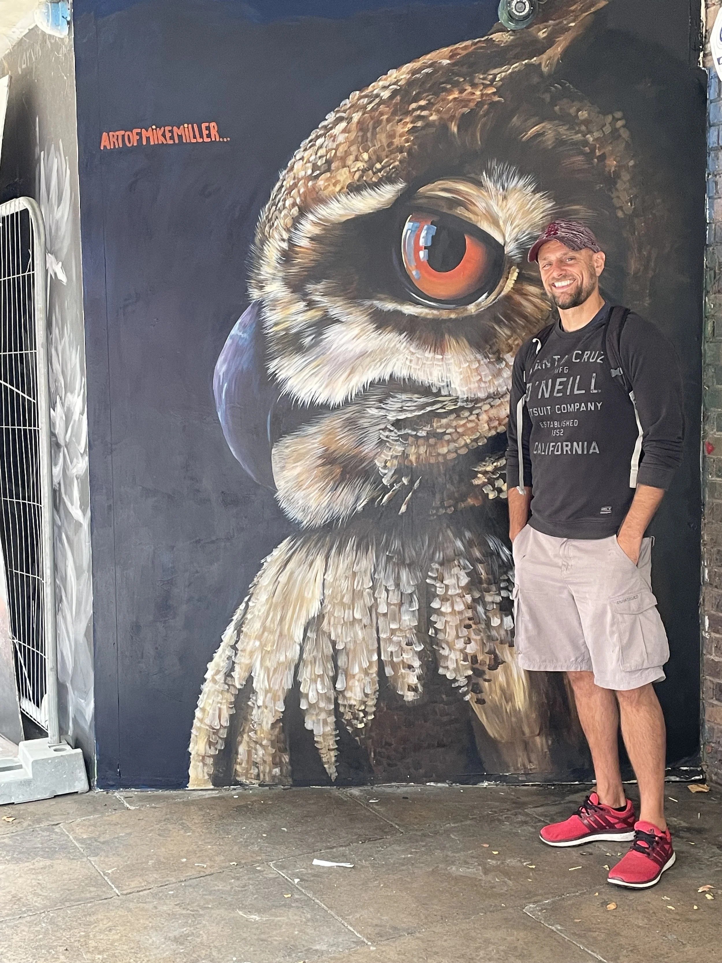A man with a beard and a baseball cap smiling while standing next to a large, realistic owl mural painted on a wall. The man is wearing a black long-sleeve shirt, beige shorts, and red athletic shoes. The owl mural depicts a close-up of the owl's eye and part of its face, with detailed feathers and beak. The background shows an urban setting with a sidewalk.