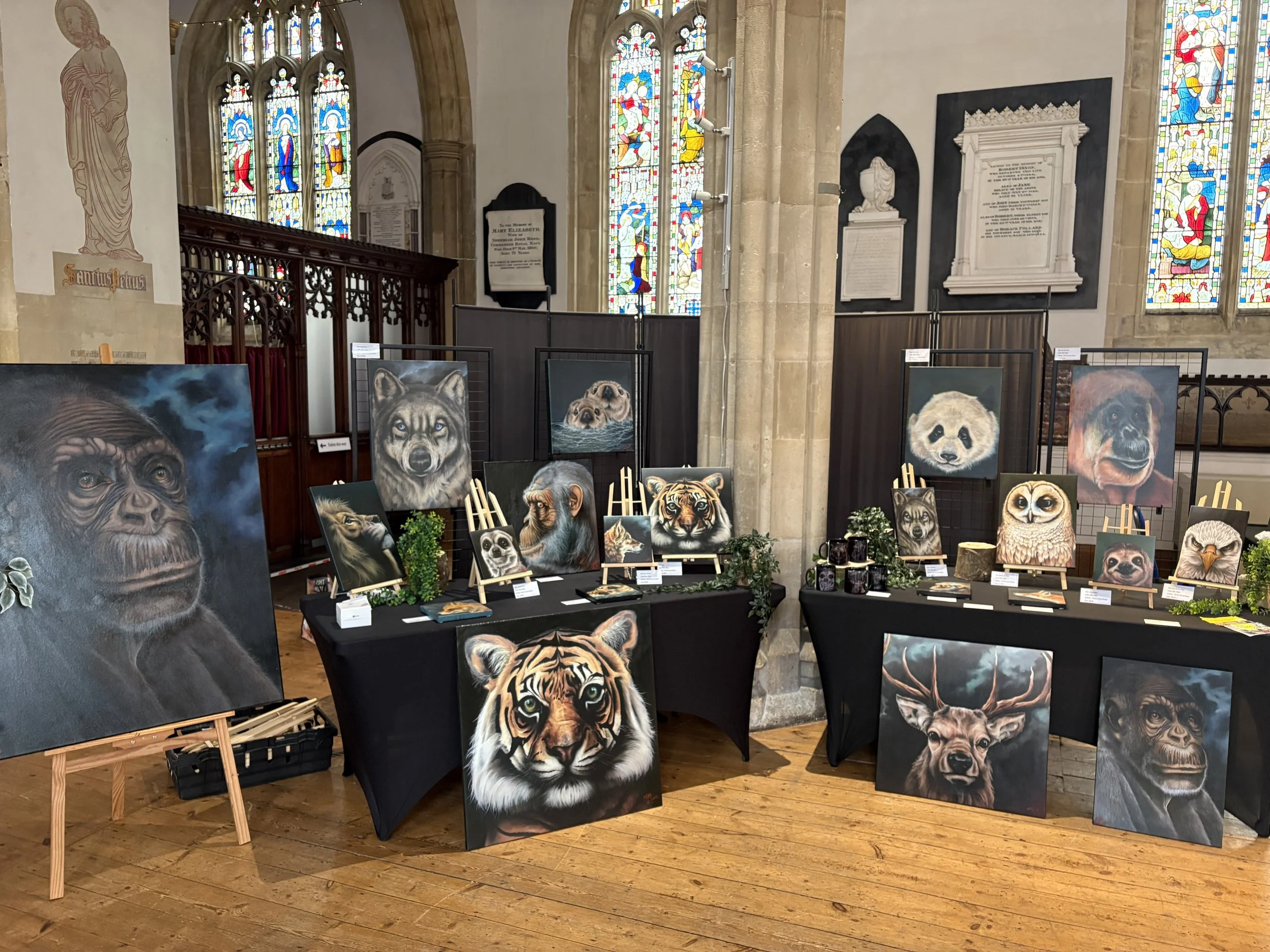 Art exhibit in a church showcasing animal paintings such as lions, wolves, pandas, bears, and deer, displayed on easels and tables with greenery, inside a church with stained glass windows.