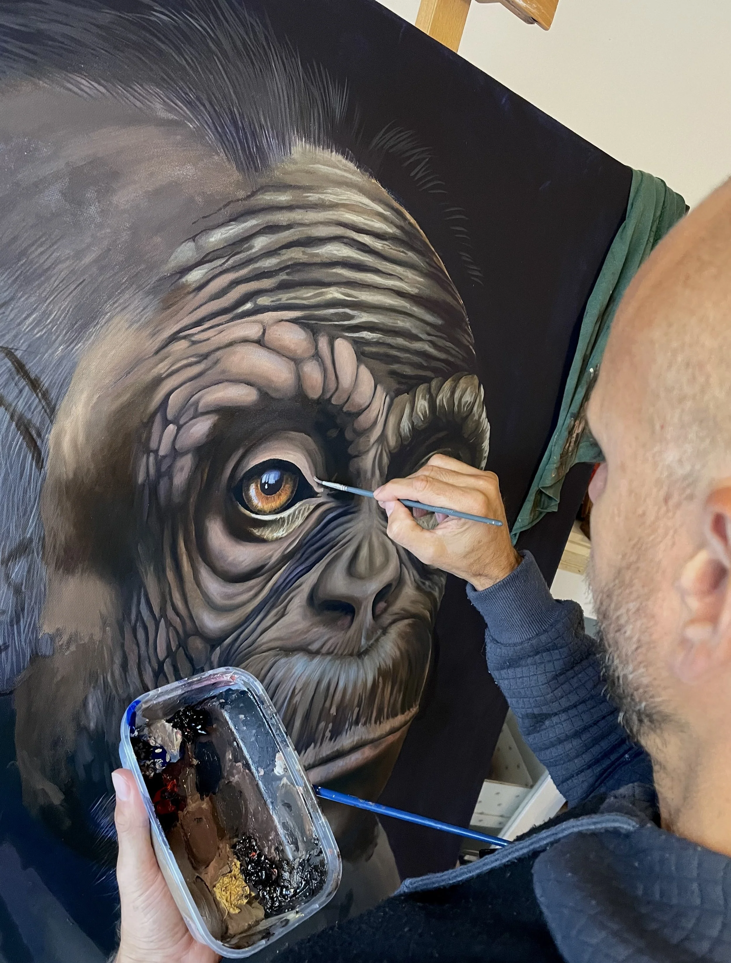 A person painting a hyper-realistic portrait of an elderly Chimpanzee with detailed wrinkles and bright eyes on canvas. The painter is seen closely working on the artwork, holding a small paintbrush.