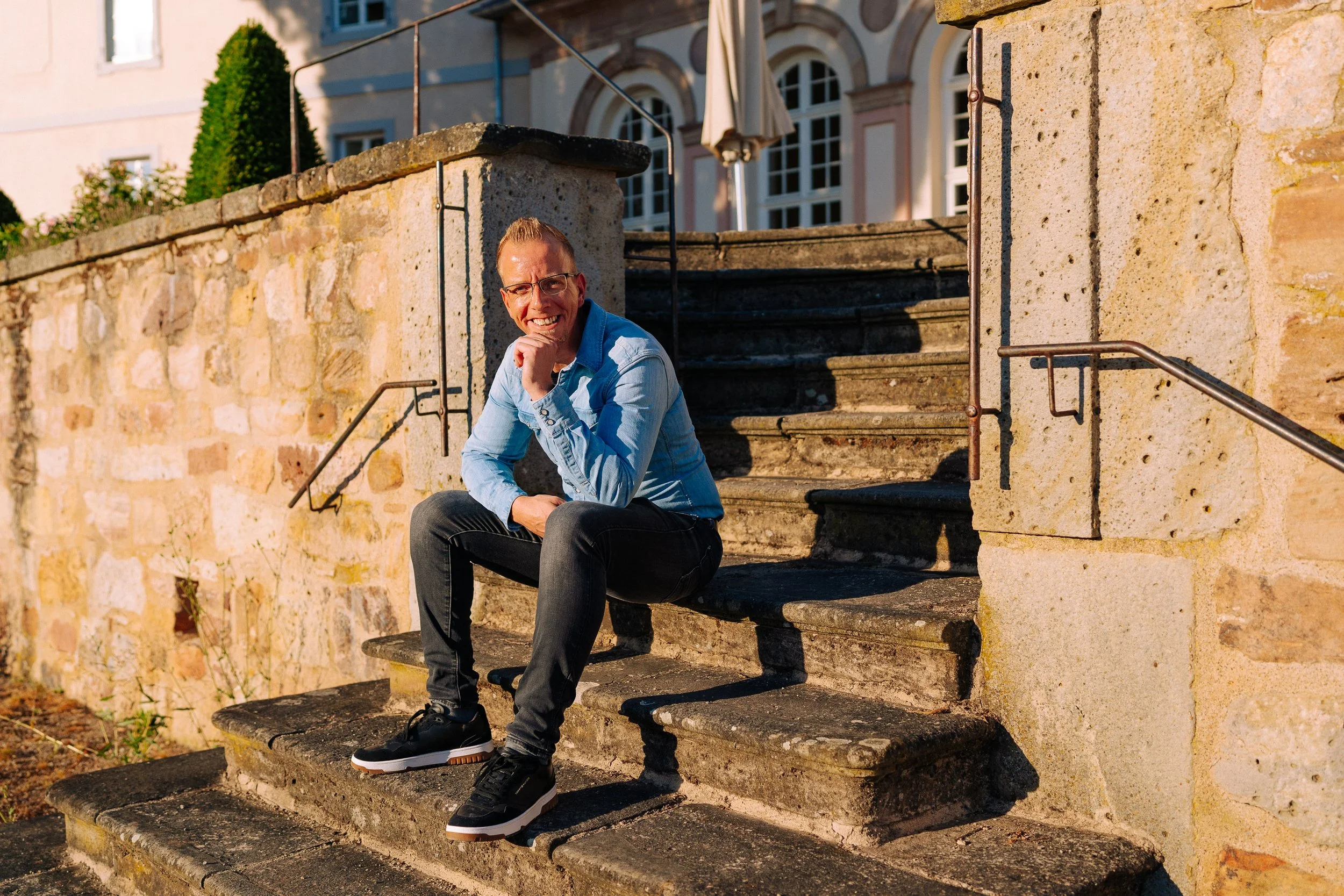 Ein Mann sitzt auf Steinblöcken an einer Außentreppe vor einem historischen Gebäude mit großen Fenstern und einem Garten im Hintergrund, die Sonne scheint.