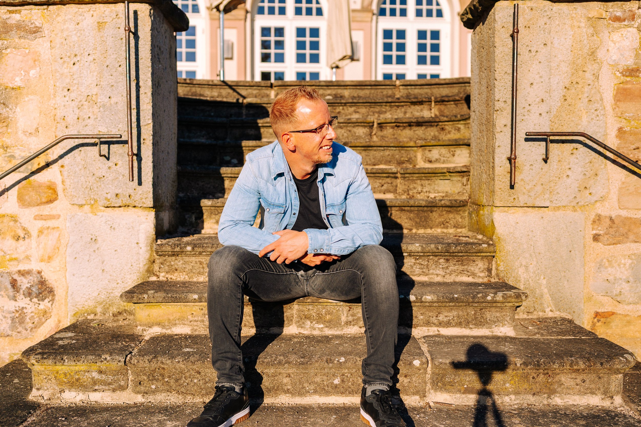 Ein junger Mann mit Brille, blauer Jeansjacke und schwarzen Schuhen sitzt auf einer alten Steintreppe vor einem historischen Gebäude mit großen Fenstern und Steinmauer, bei Sonnenschein.