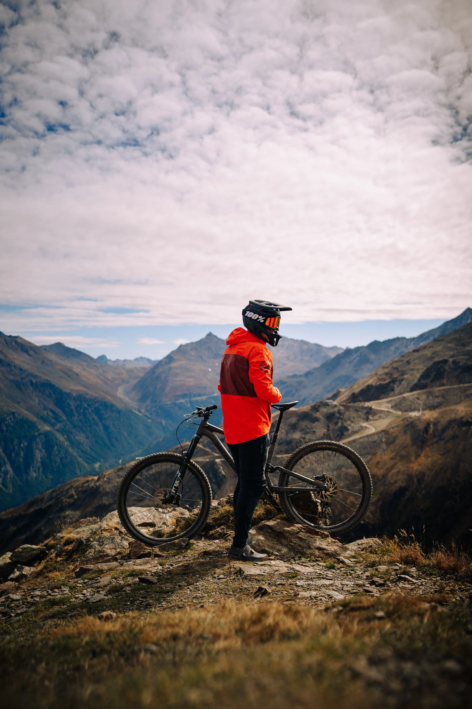 Ein Mountainbiker in einem roten Jacke und schwarzer Hose steht mit seinem Mountainbike an einem felsigen Wanderweg in einer Berglandschaft mit hohen Bergen im Hintergrund. Der Himmel ist überfüllt mit Wolken.