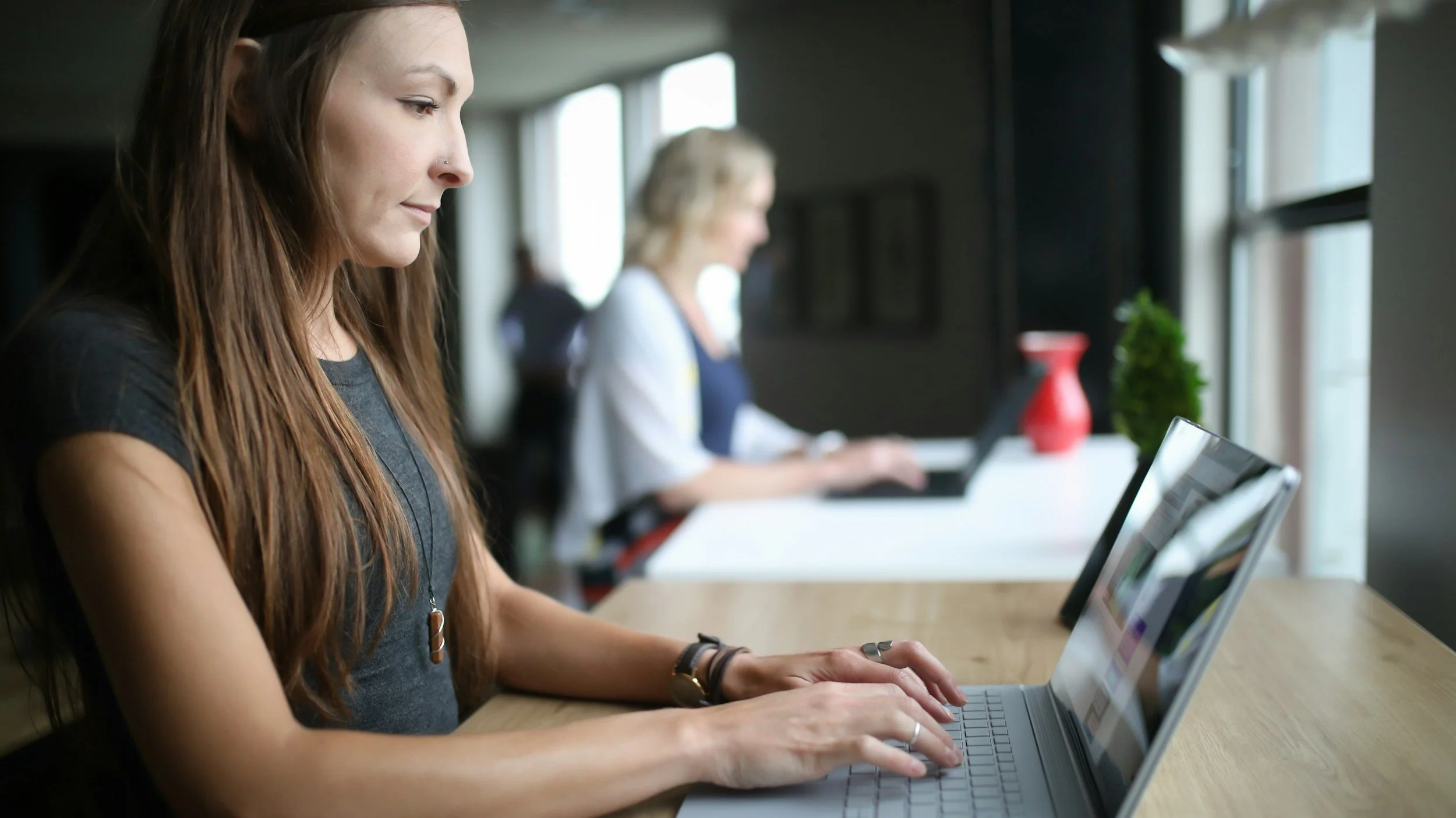 A young woman with long brown hair working on a silver laptop in a modern office, with other people working on laptops in the background.