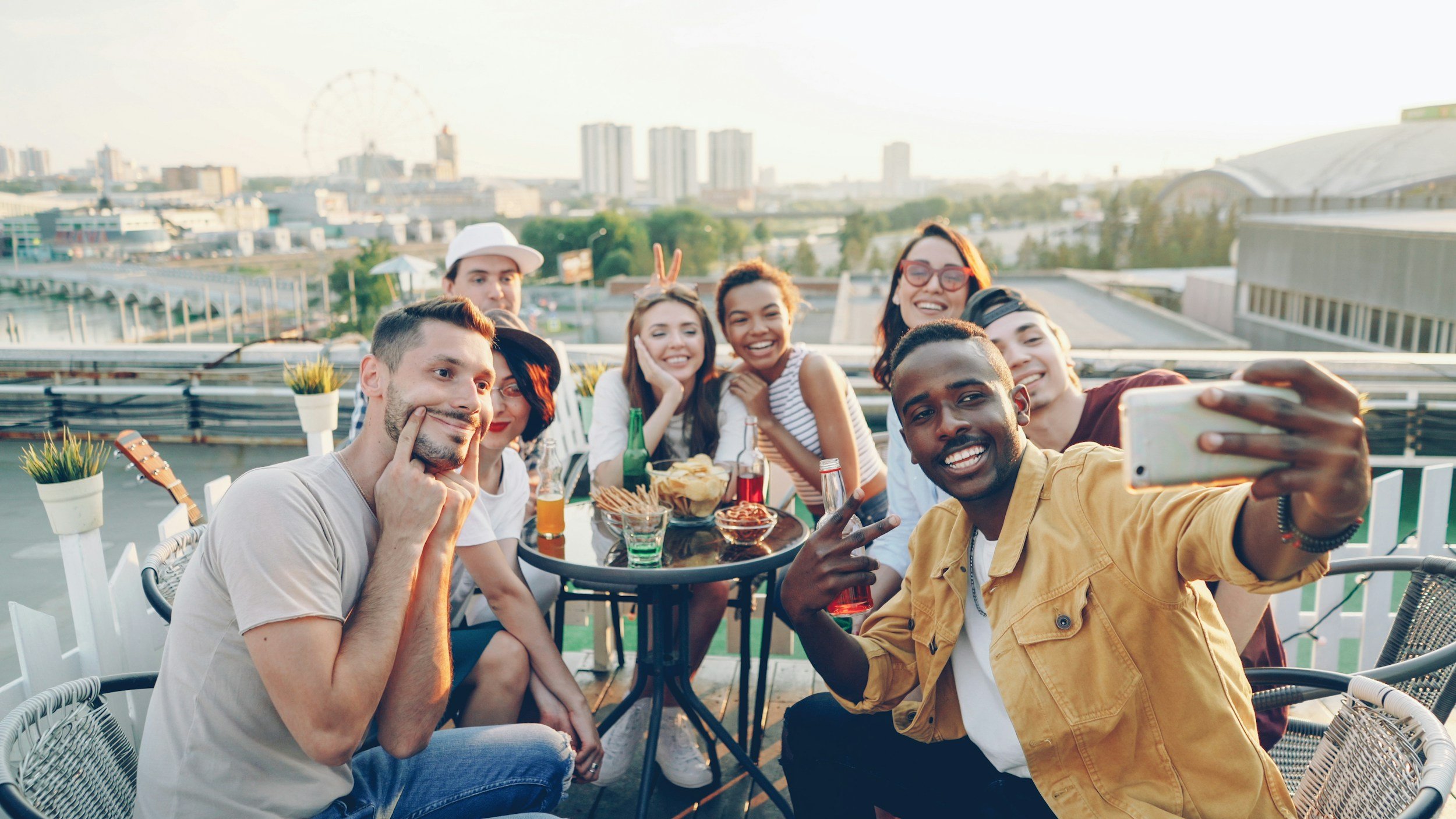 A group of friends taking a selfie on a rooftop terrace with city buildings in the background, enjoying food and drinks during sunset.