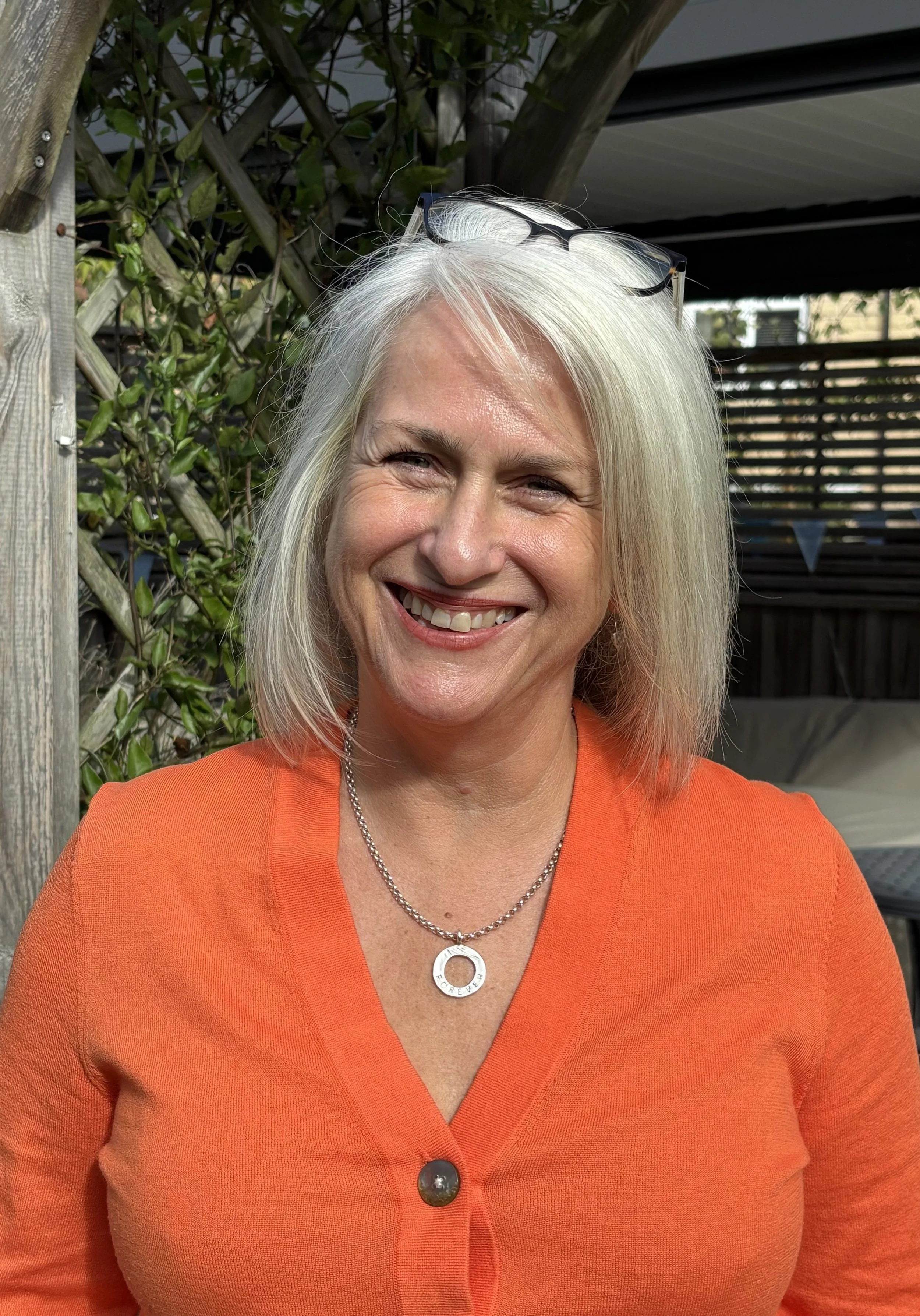 A woman with platinum blonde hair smiling, wearing an orange top, glasses on her head, and a silver necklace with a circular pendant that says 'Forever'.
