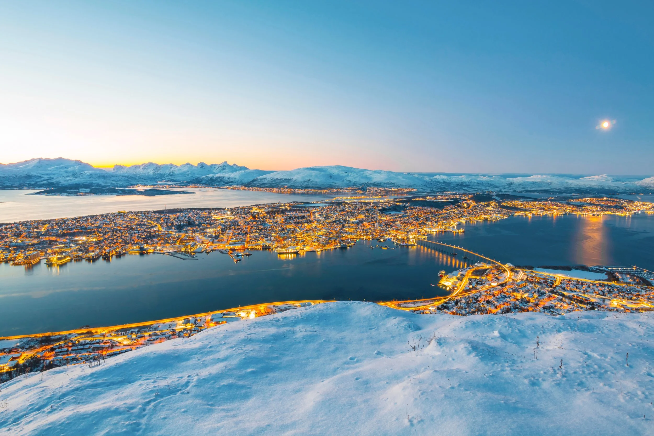 Aerial view of a city by a lake with illuminated streets and snow-covered mountains in the background during twilight.