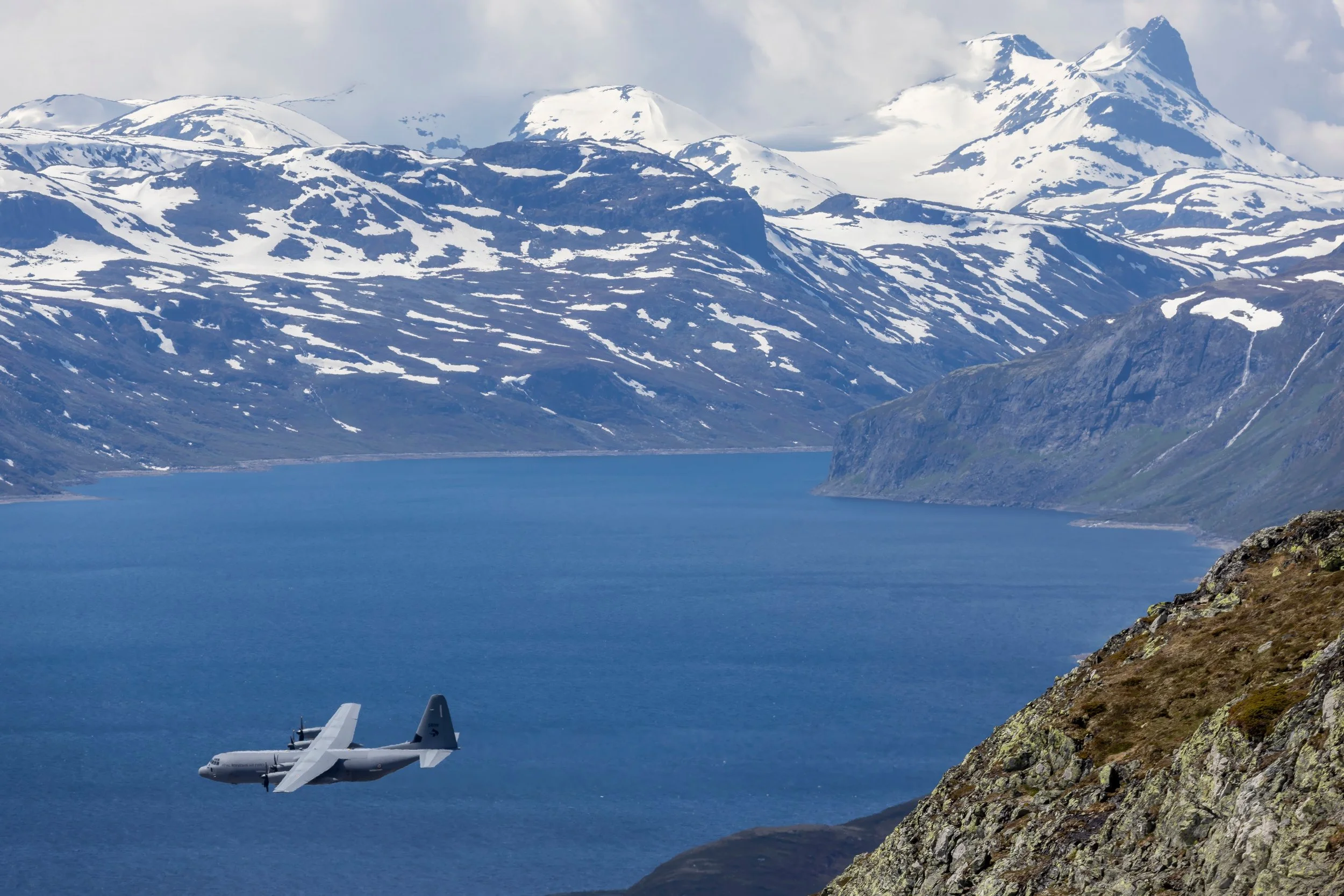 a military aircraft flying over a body of water with snow-covered mountains in the background
