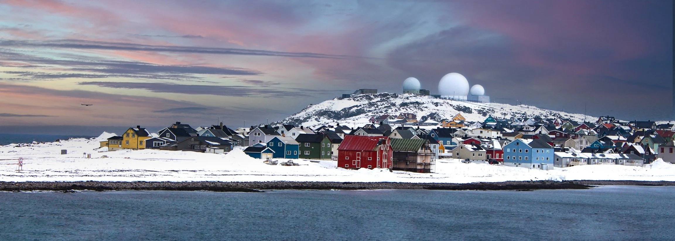 Colorful houses along a snowy shoreline with a hill topped by large white radar domes and a sunset sky in the background.