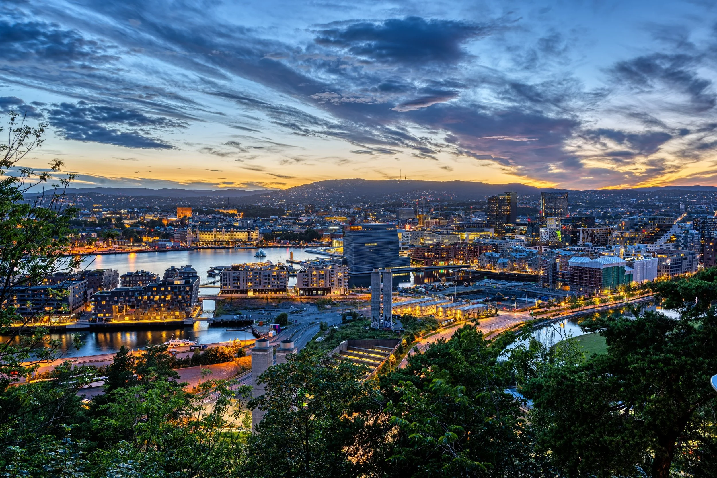 A city skyline at dusk with buildings illuminated and a river in the foreground, mountains in the background, and a partly cloudy sky with the sun setting.