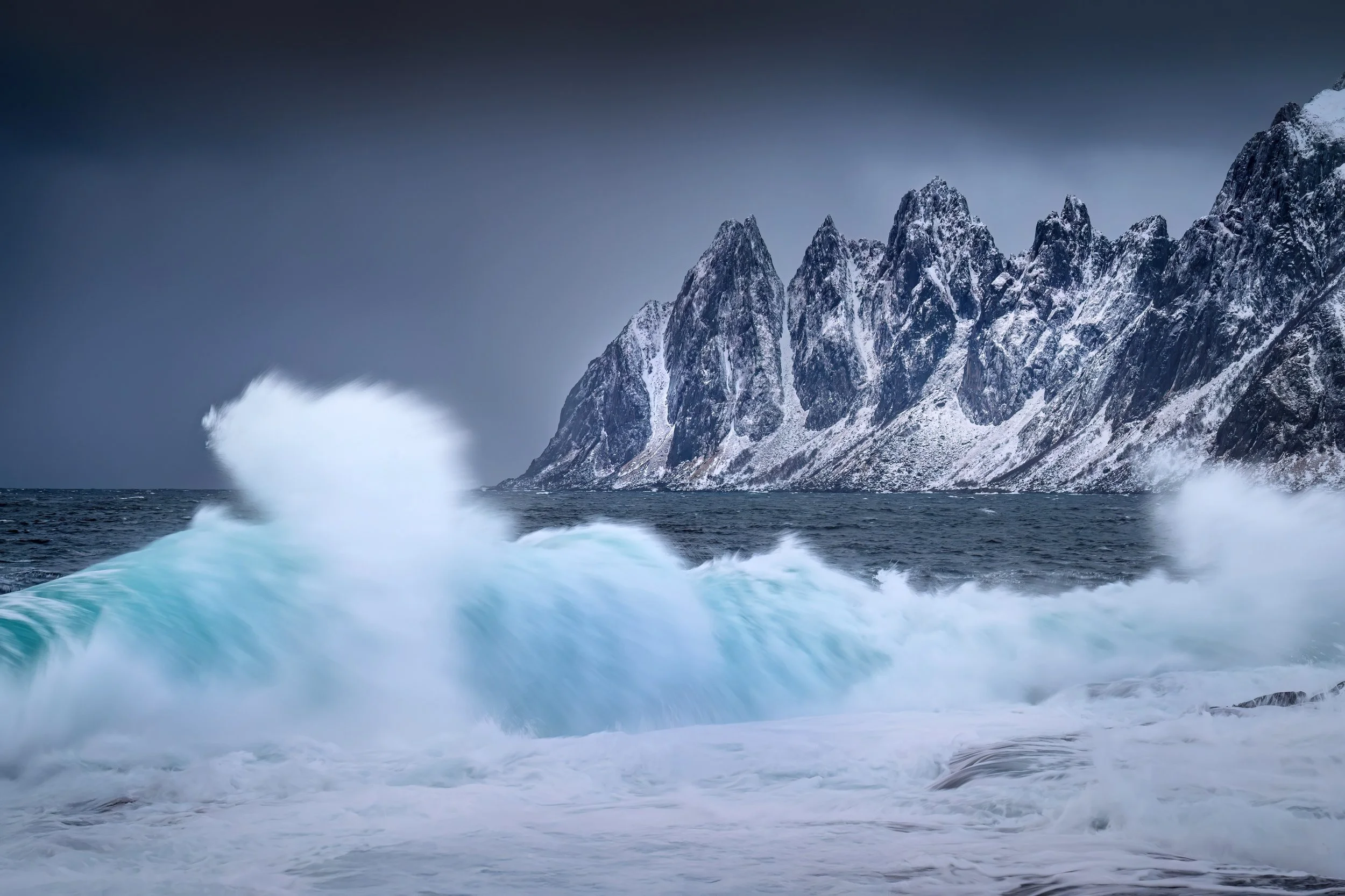 Snow-capped mountains and rocky cliffs along a rough ocean shoreline with waves crashing.