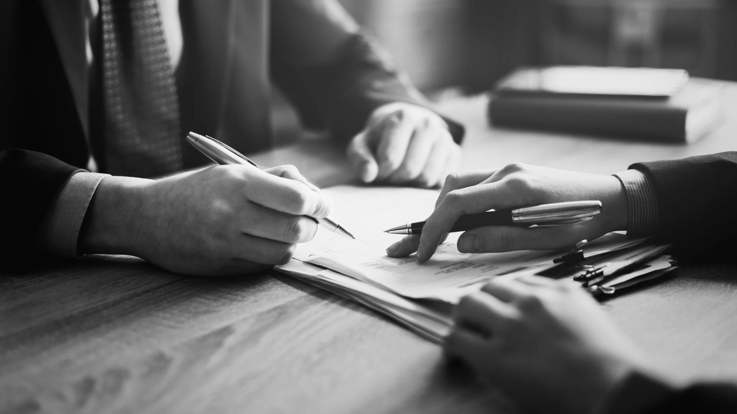 Two people sitting at a desk, engaging in a business discussion. One person is signing a document while the other's hand is pointing at the paper. Both are dressed in business attire, with a clipboard and additional papers on the desk.