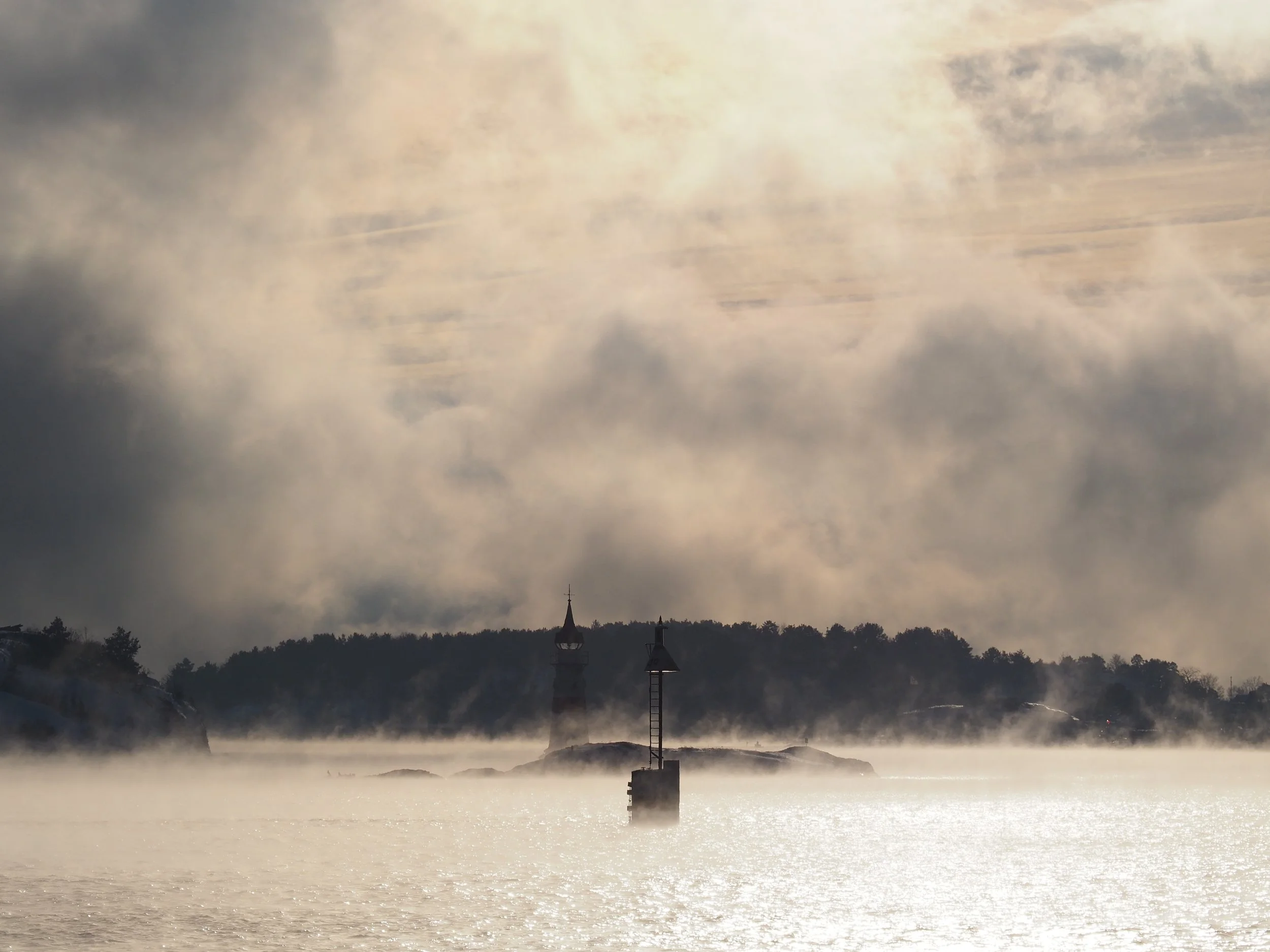 A lighthouse on a small island surrounded by water, with hills and trees in the background, under a cloudy sky.