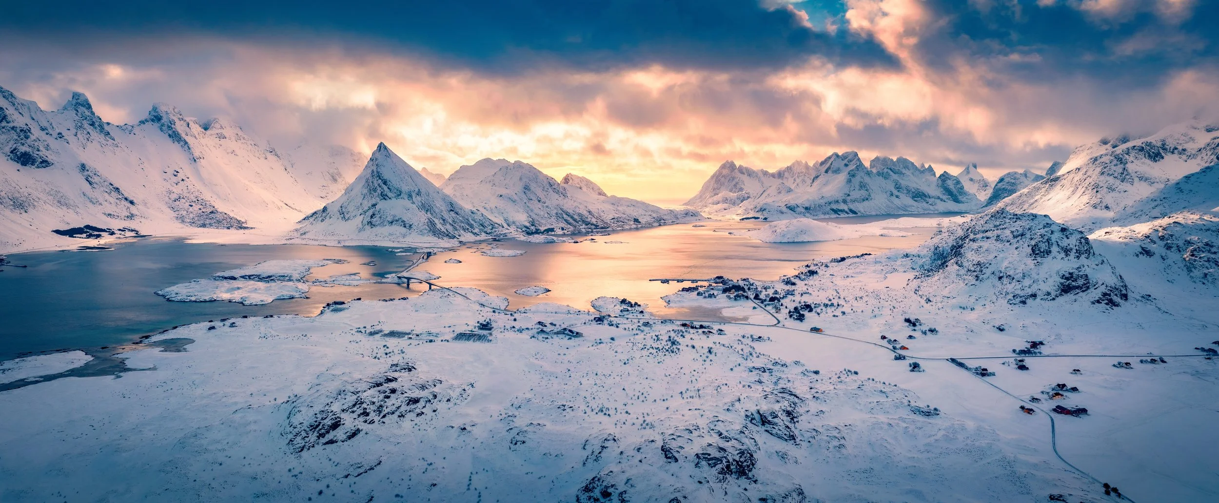 Snow-covered mountains surrounding a calm, partially frozen body of water during sunset, with a few scattered houses and roads.
