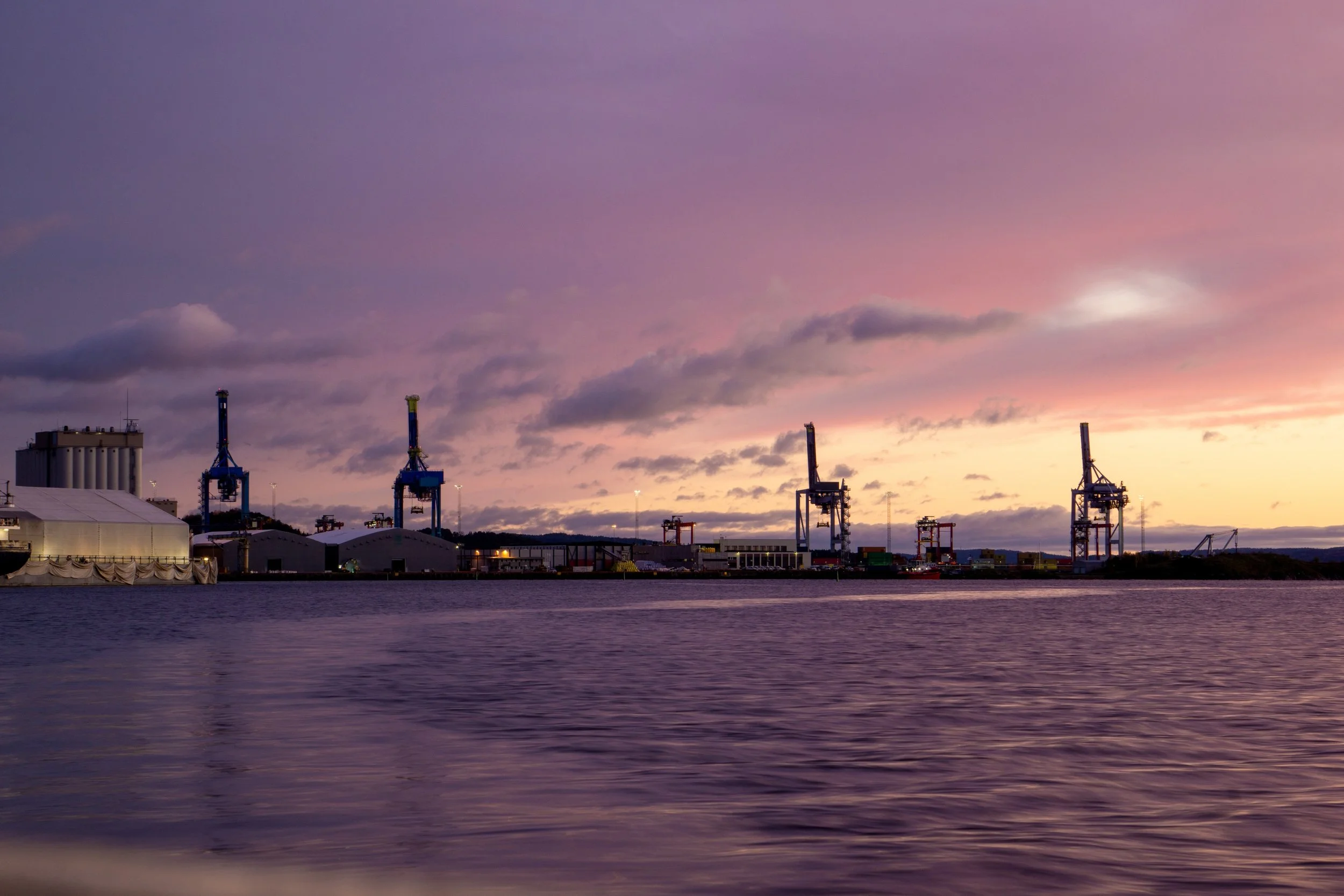 Industrial port with cranes and large warehouse buildings near water at sunset.